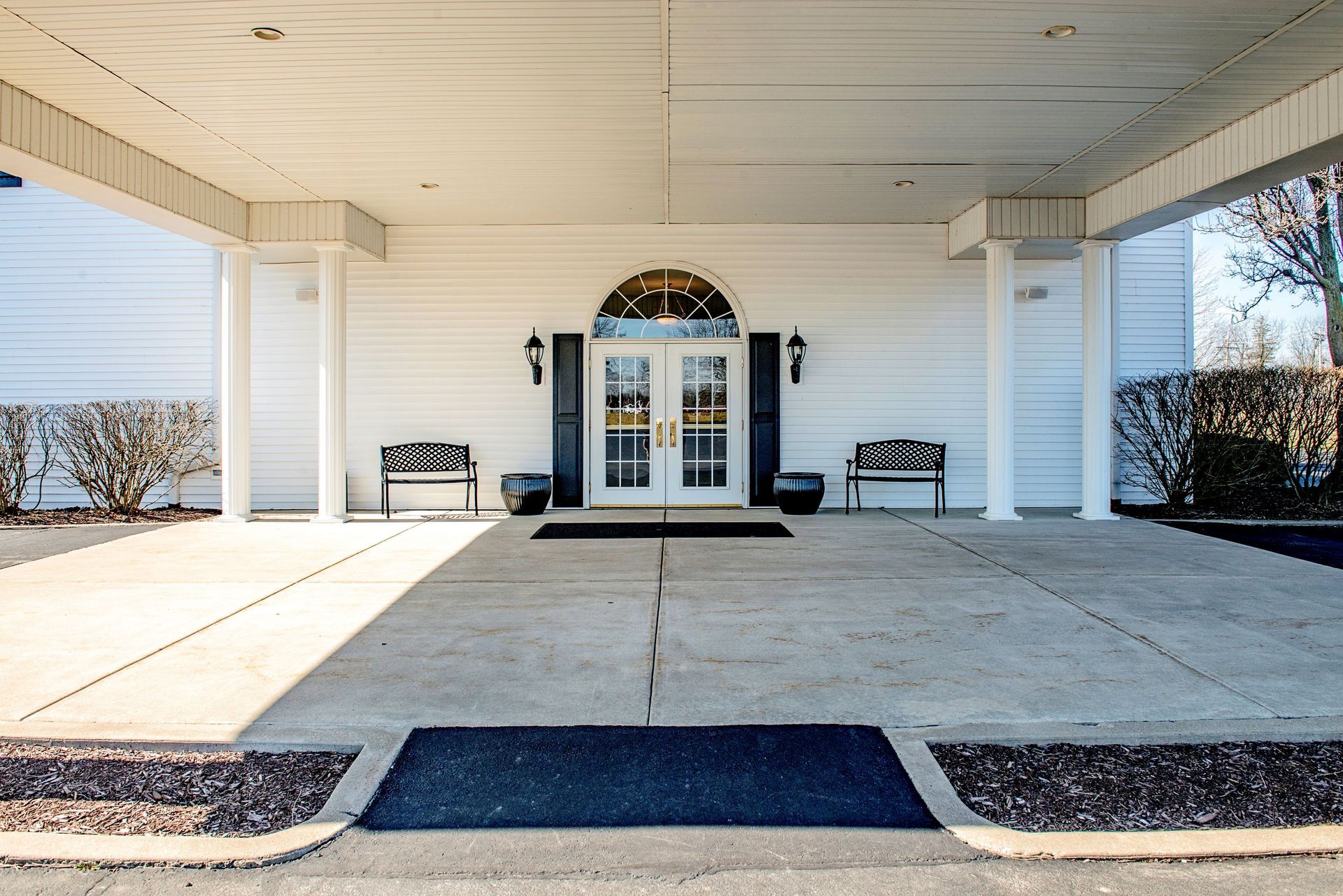 White building entrance with covered walkway, double doors, benches, and arched window.