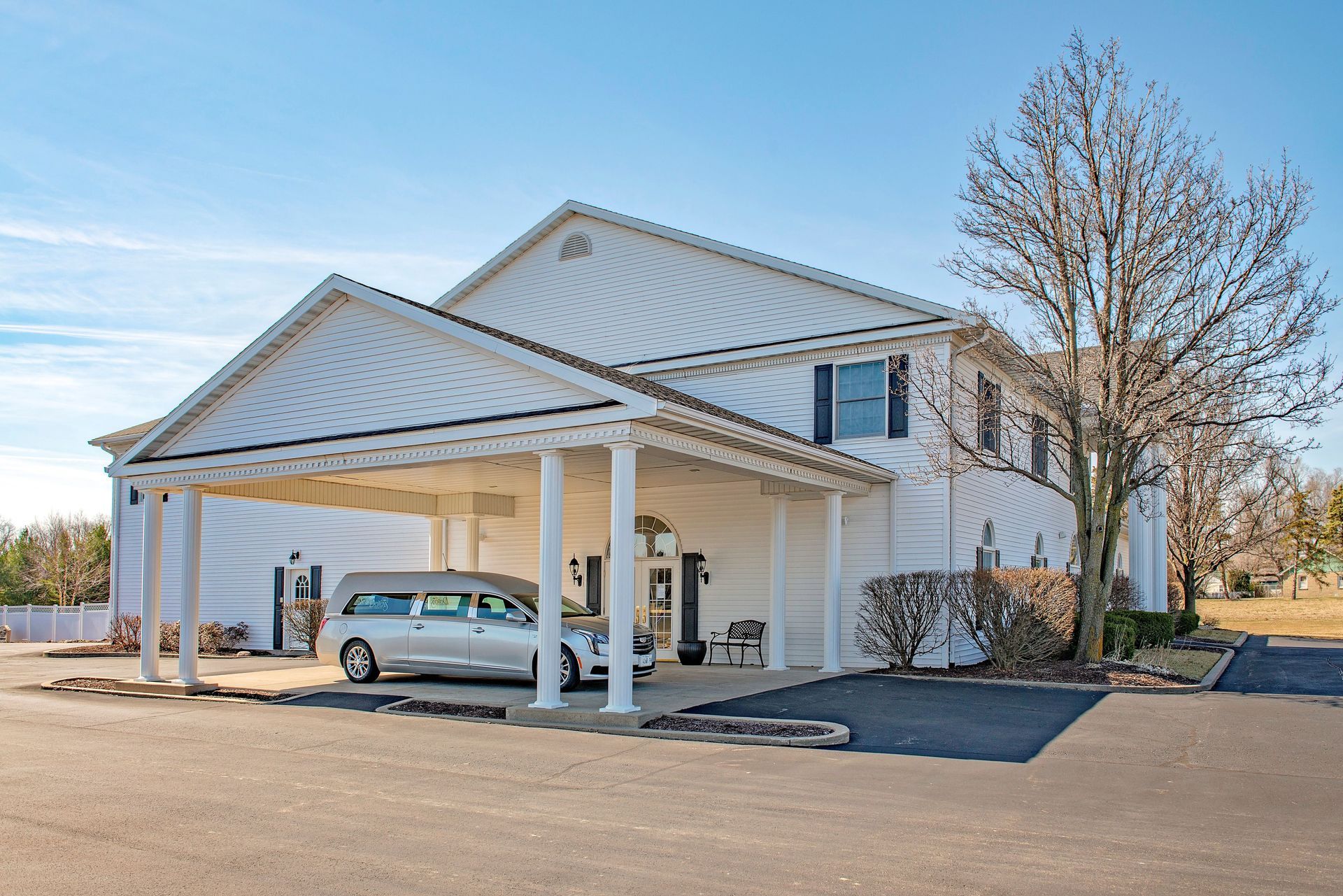 White funeral home with a hearse under a covered entrance, parked on a paved lot.