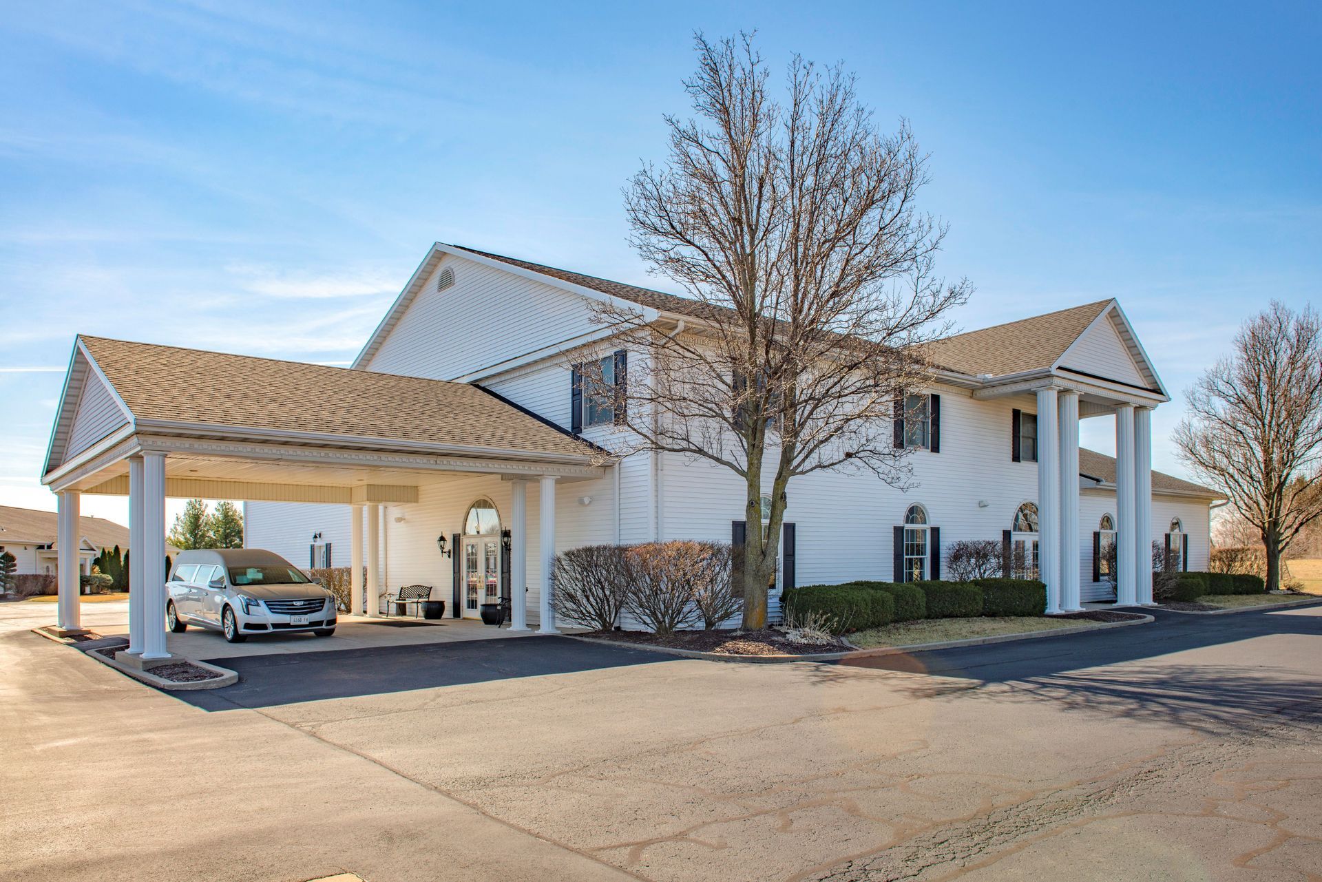 White funeral home with a porte-cochere and a parked hearse on a sunny day.