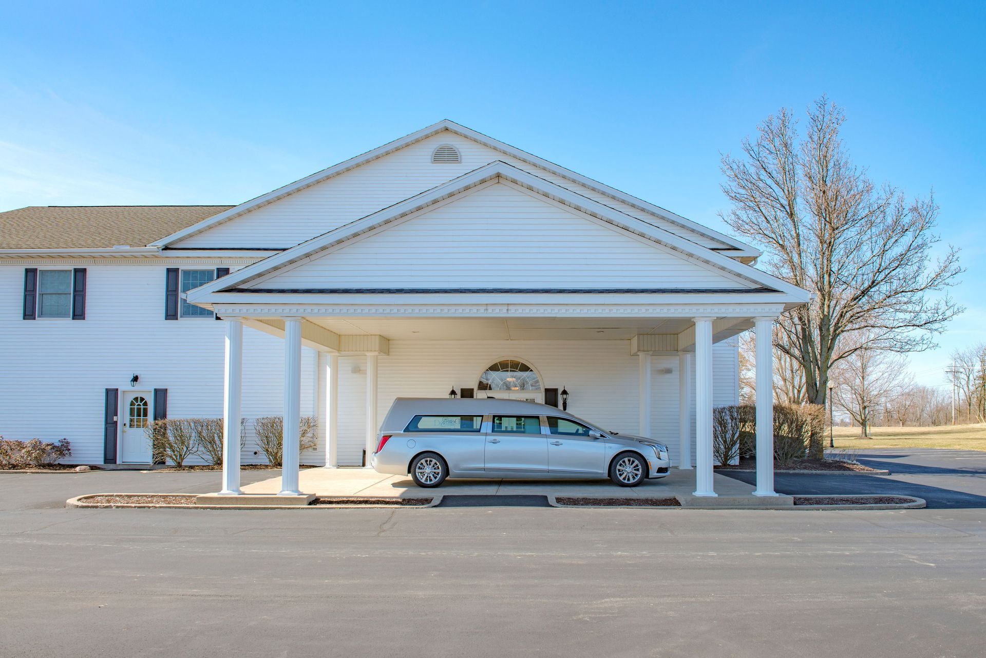 White hearse parked under a white covered entrance of a funeral home on a sunny day.