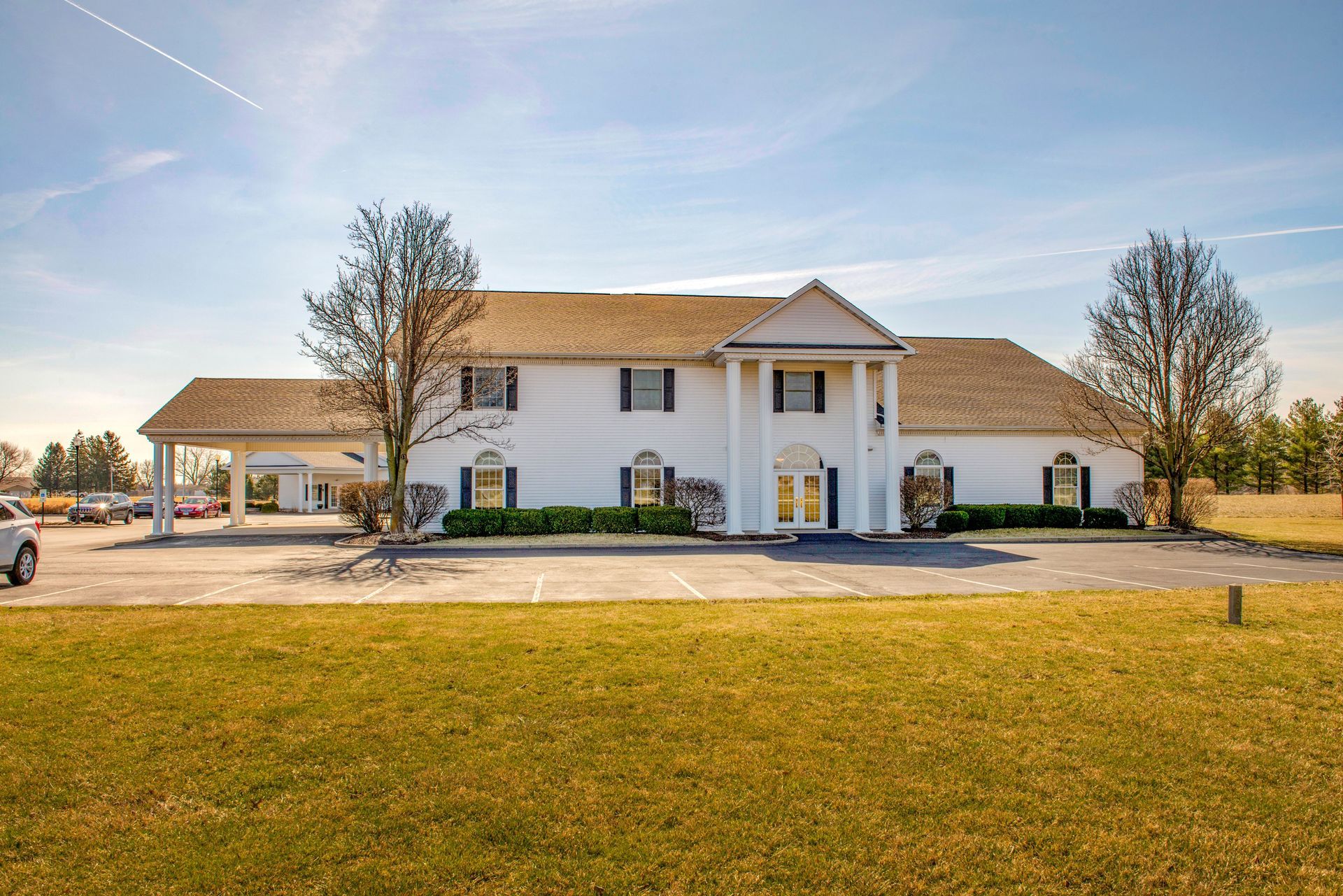 White building with pillars, likely a funeral home, on a sunny day with green grass and parking.
