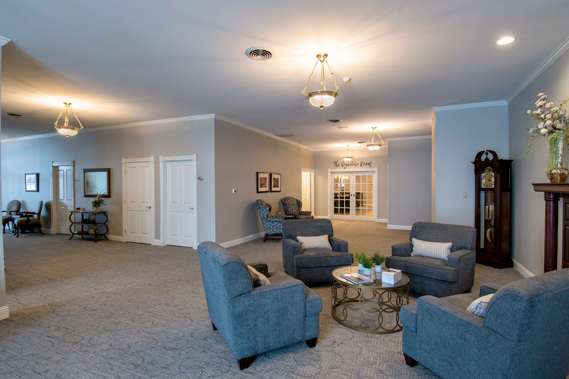 Lounge area with blue armchairs, round coffee table, gray carpet, and chandeliers in a light-filled room.