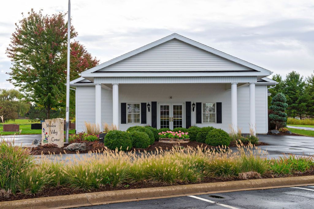White house with porch and columns beside wet road, framed by trees and shrubs