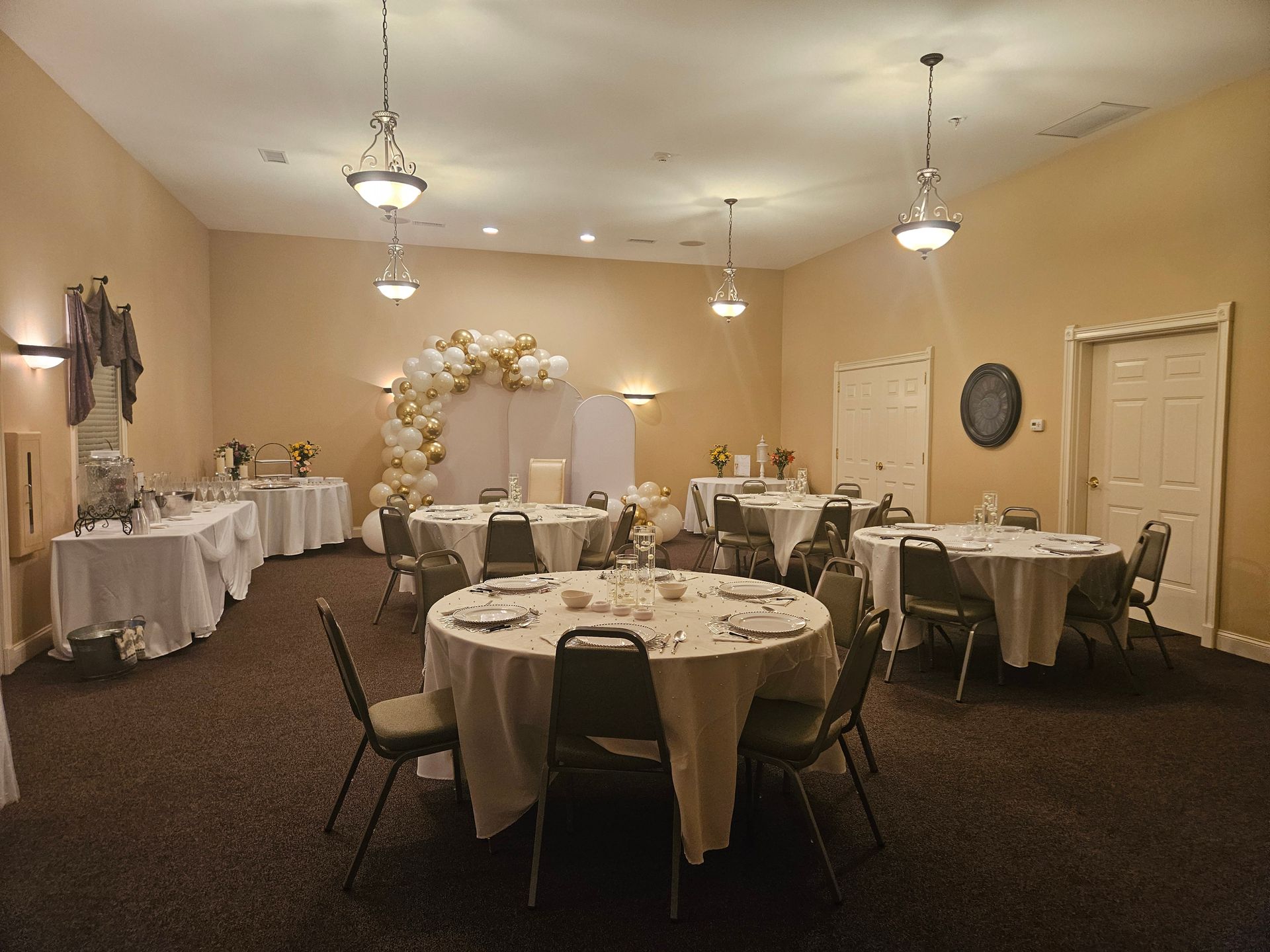Decorated banquet room with round tables, white tablecloths, chairs, and a balloon arch near the back wall