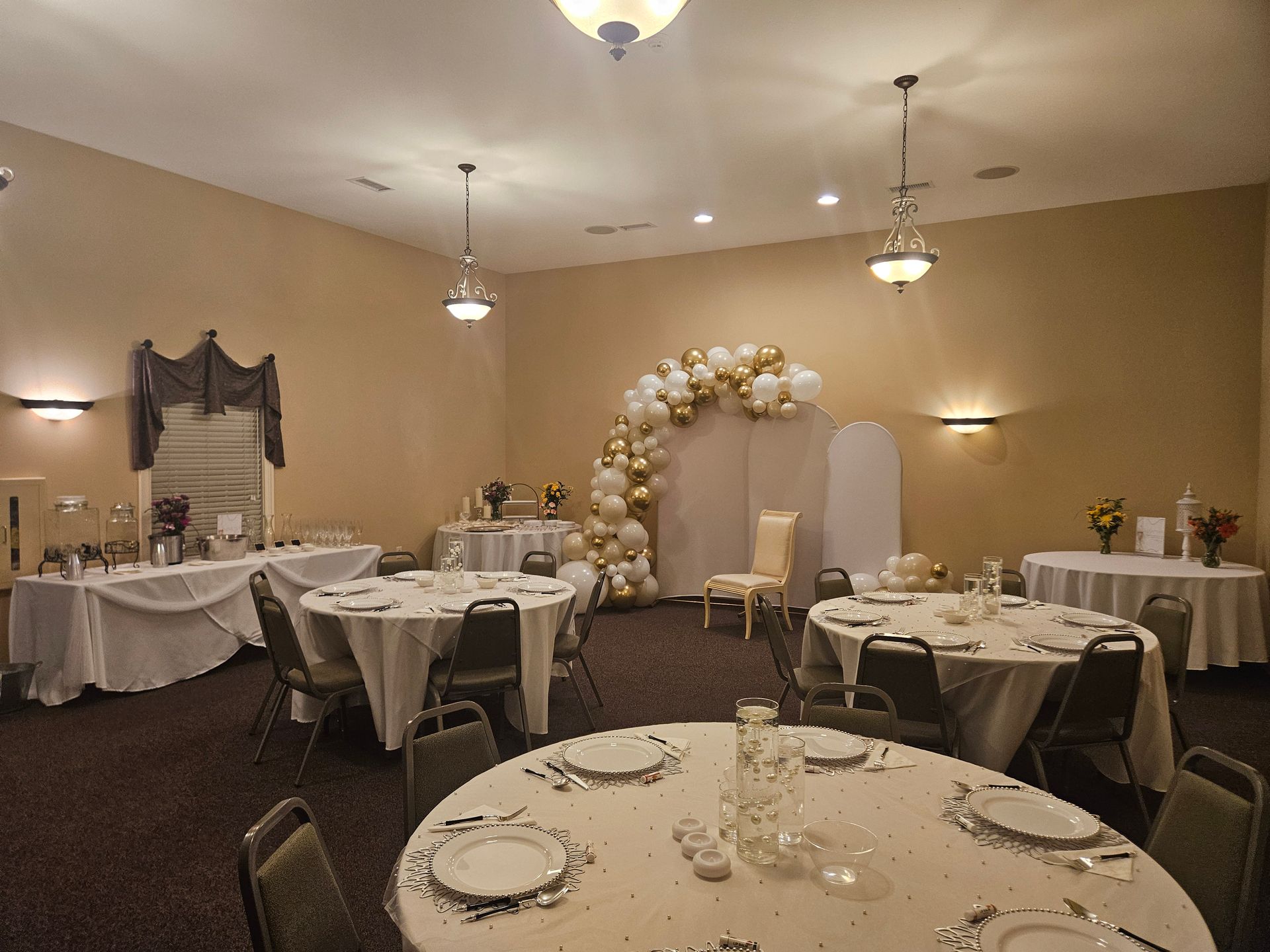 Decorated banquet hall with round tables, white linens, and a gold-and-white balloon arch near the back wall