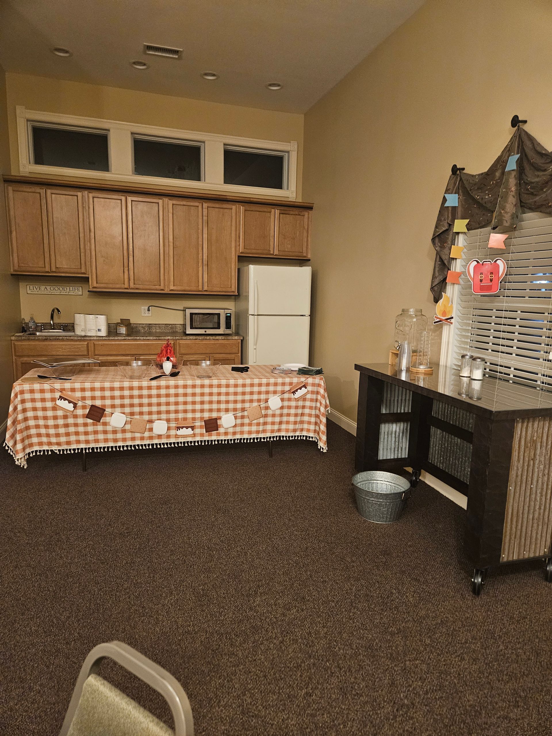 Community room with kitchen counter, white refrigerator, checkered table, and shelving unit in a carpeted space