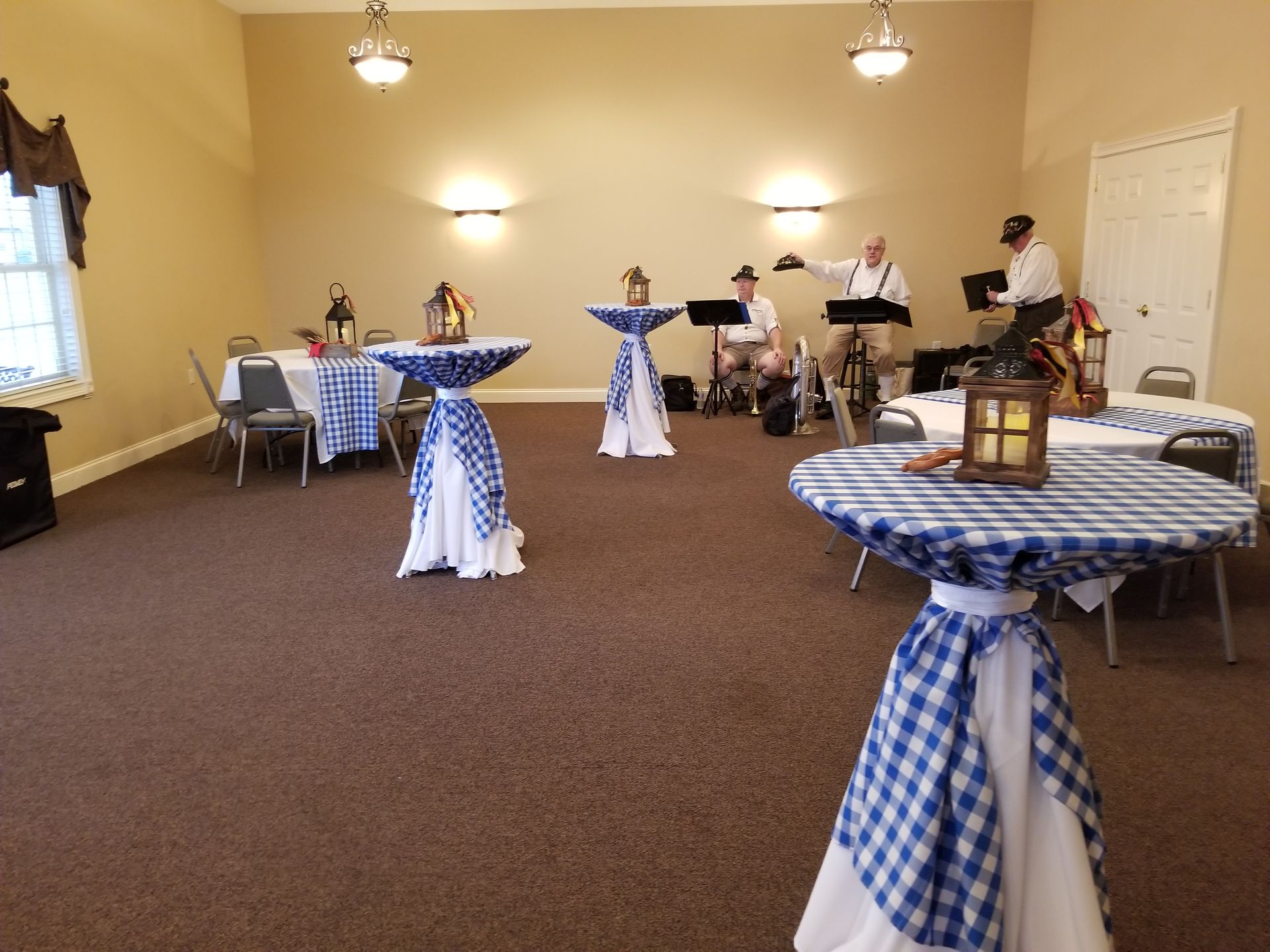 Banquet room with blue gingham-covered tables, lantern centerpieces, and people setting up in the background