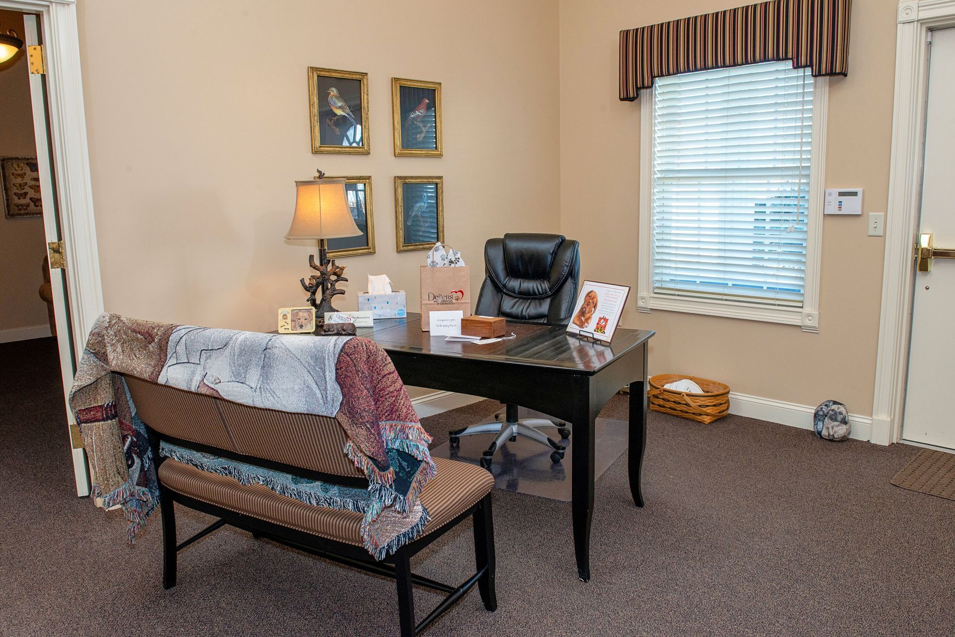 Office with desk, black chair, bench with blanket, framed art, and window with blinds.