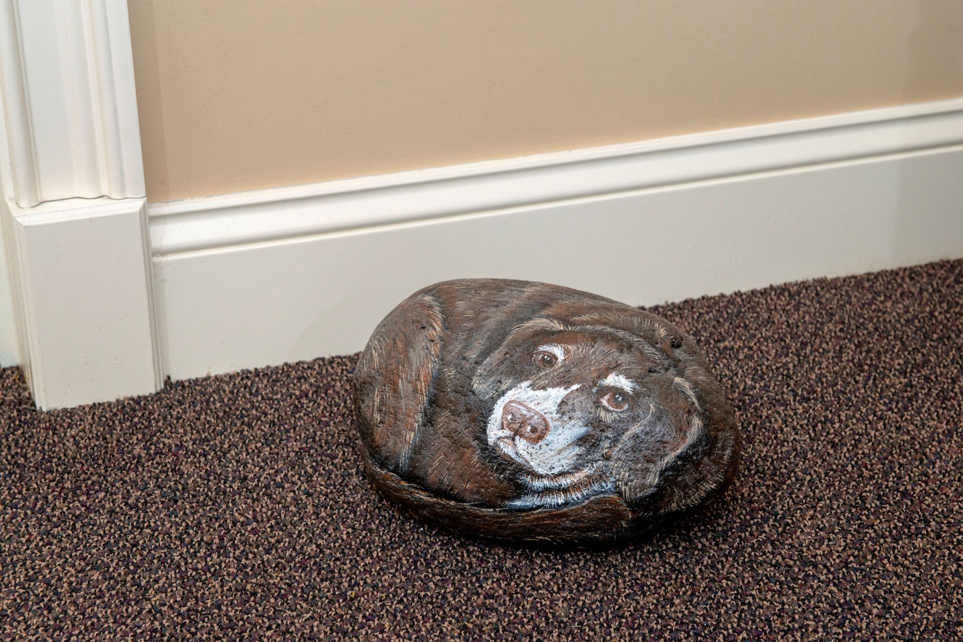 Rock painted with dog's face, on brown carpet next to a beige wall with white trim.