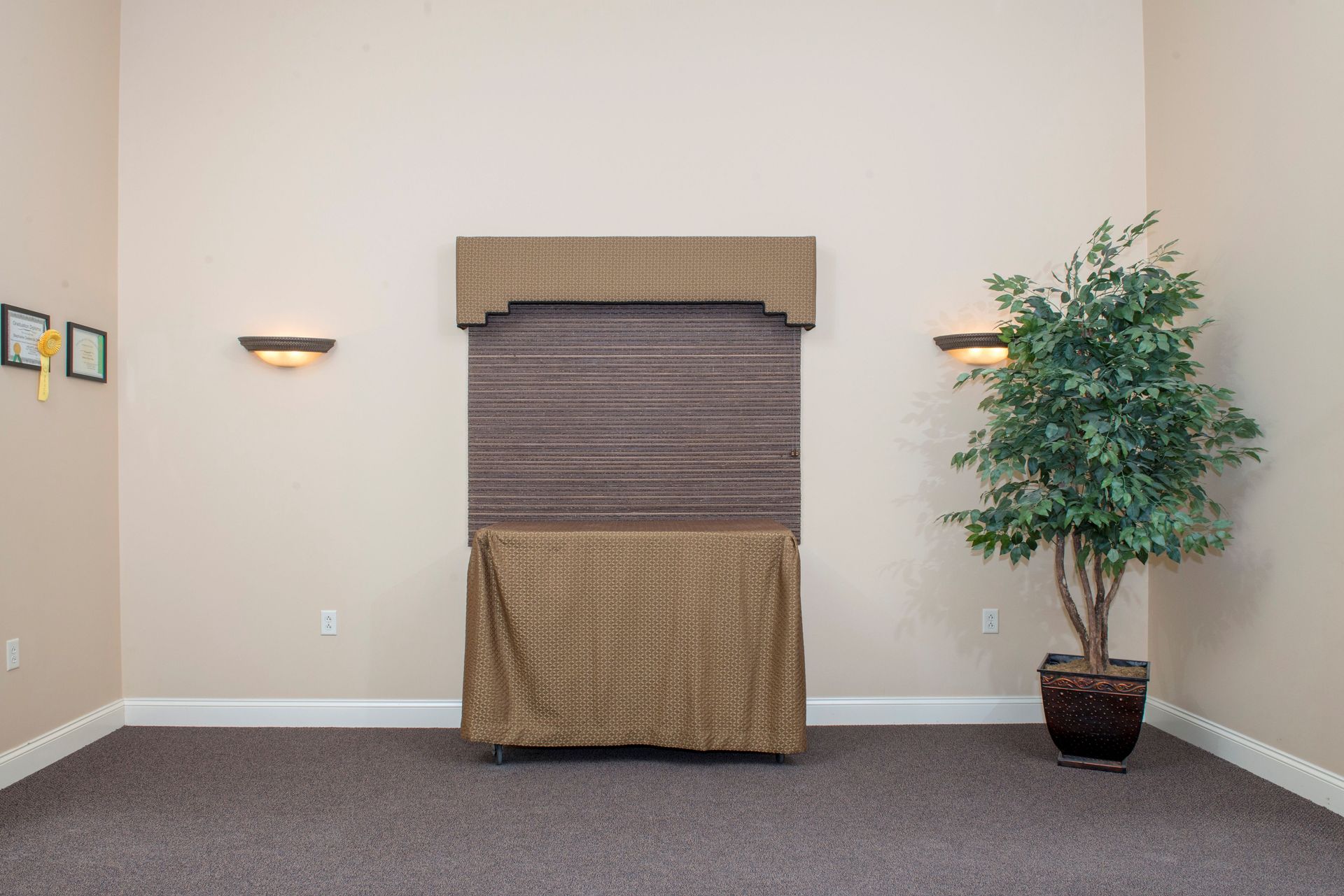 A large brown display with a patterned drape, flanked by wall sconces and a potted tree, in a beige room.