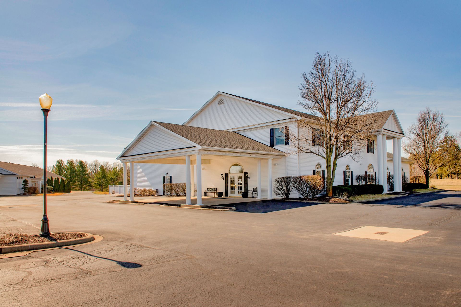 White two-story building with a covered entrance, likely a funeral home, on a paved lot under a blue sky.