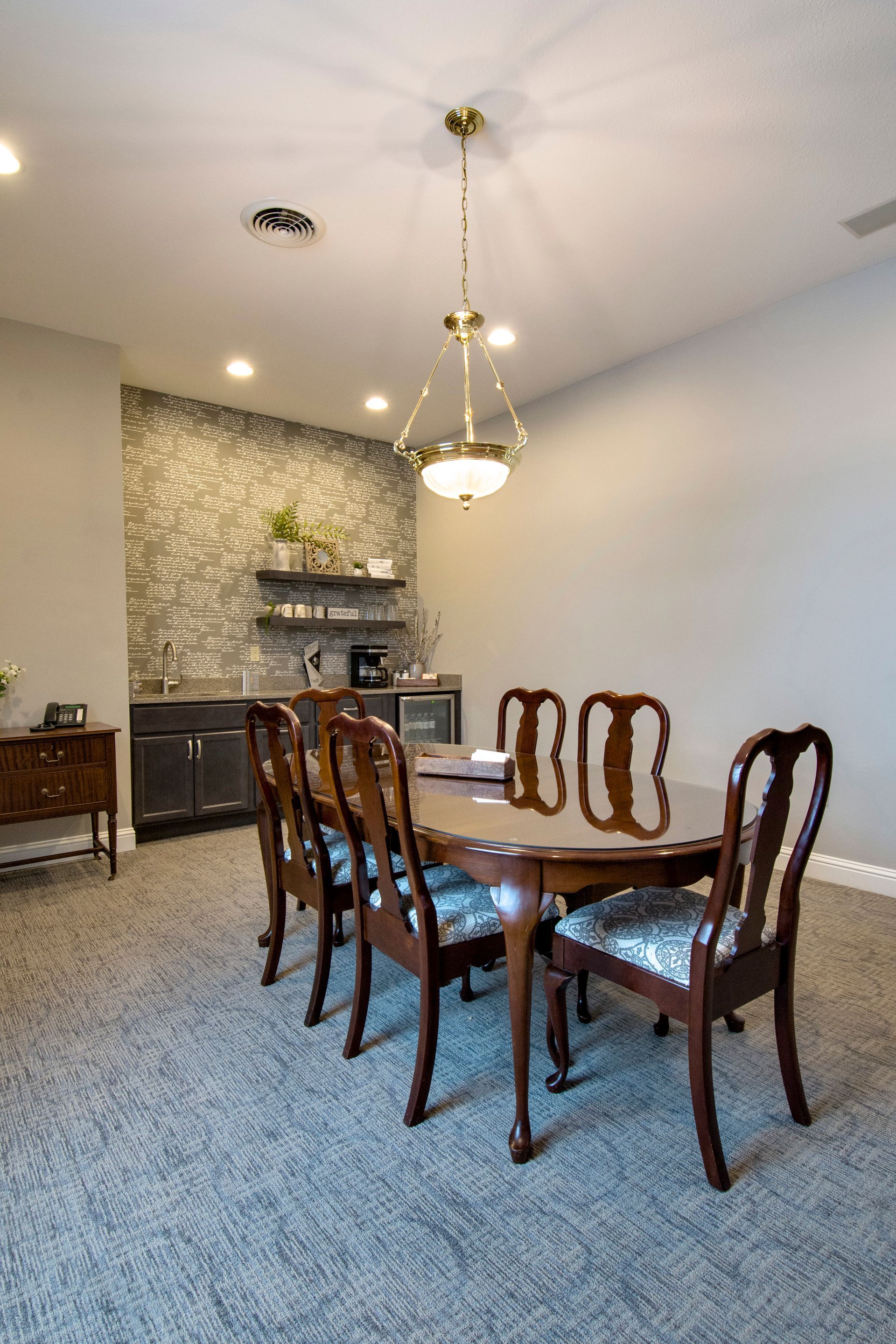Dining room with oval wooden table, six chairs, built-in bar, and chandelier. Gray carpet and walls.