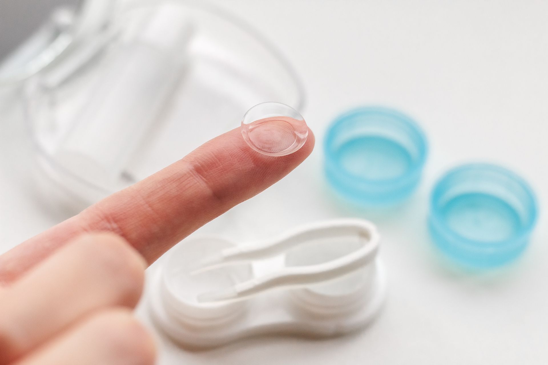 Finger hält durchsichtige Kontaktlinse neben Linsenbehälter und blauen Lösungsdeckeln