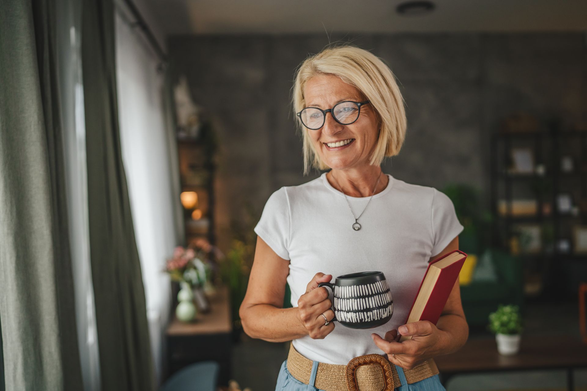 Eine lächelnde Person hält eine Tasse und ein Buch in einer gemütlichen Inneneinrichtung.