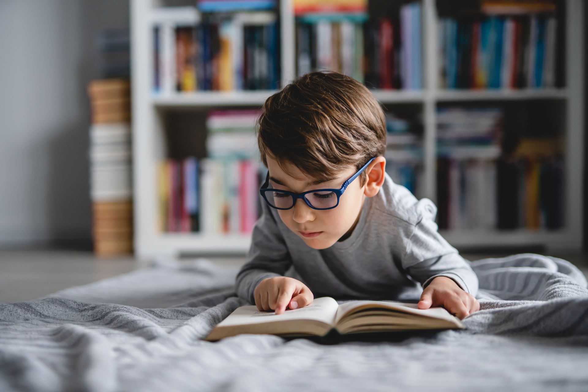 Junge liest auf dem Bett ein Buch, trägt eine blaue Brille in einem gemütlichen Schlafzimmer