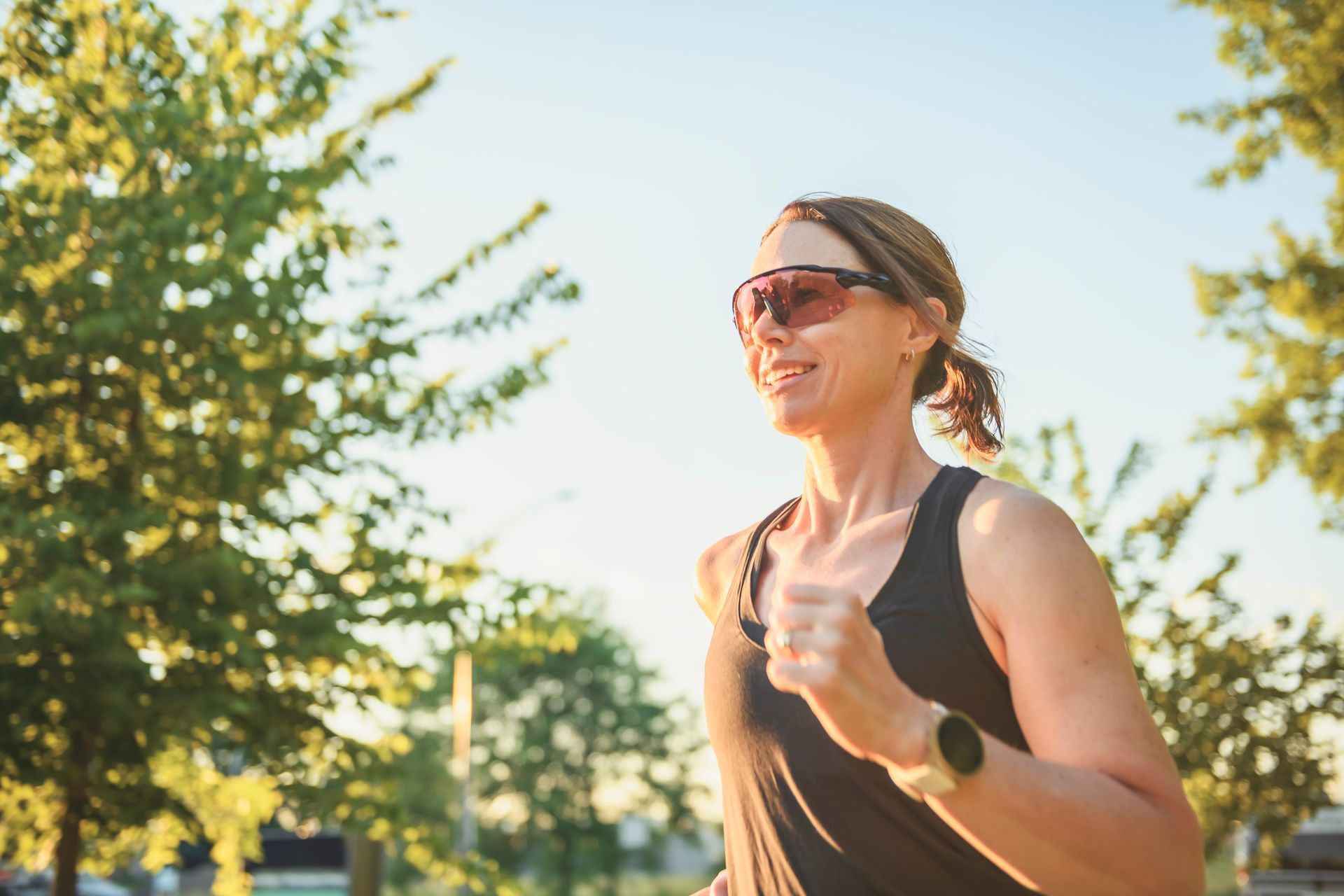 Eine Frau joggt im Freien im Sonnenlicht; sie trägt eine Sonnenbrille und ein schwarzes Tanktop.