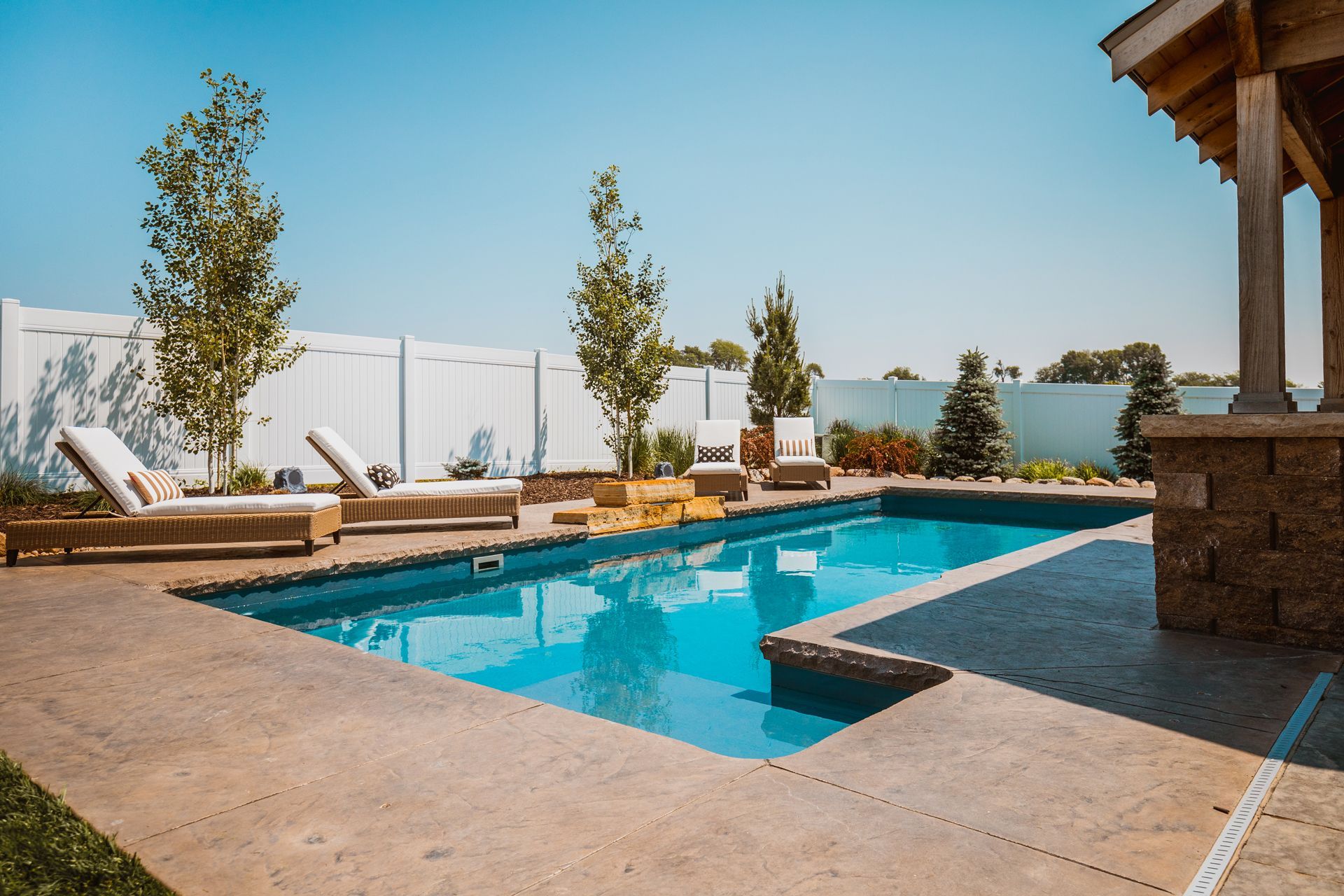 Rectangular pool in a backyard with lounge chairs, trees, and a gazebo on a sunny day.