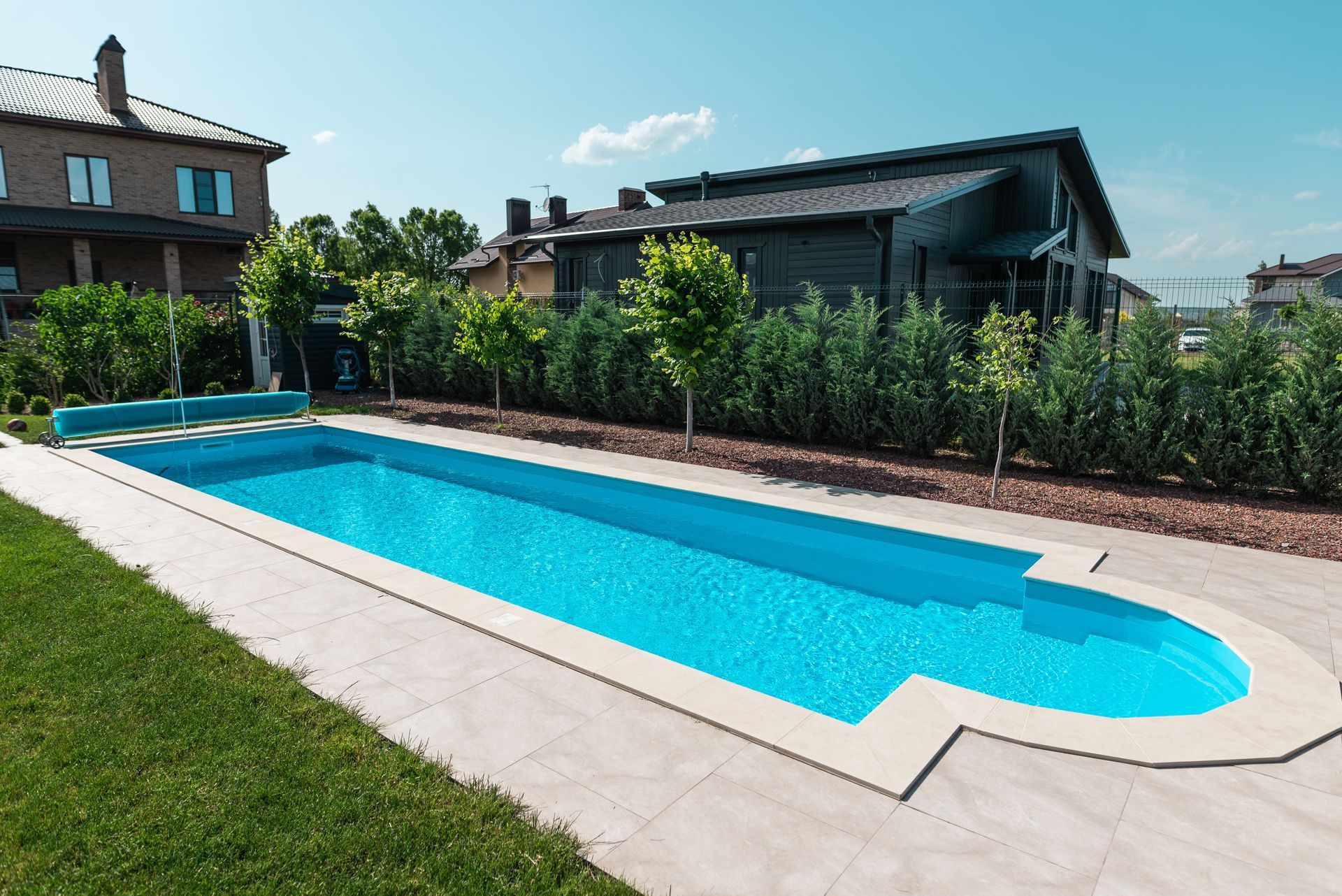 Long, rectangular pool with blue water and surrounding stone patio, in front of houses and green hedges.