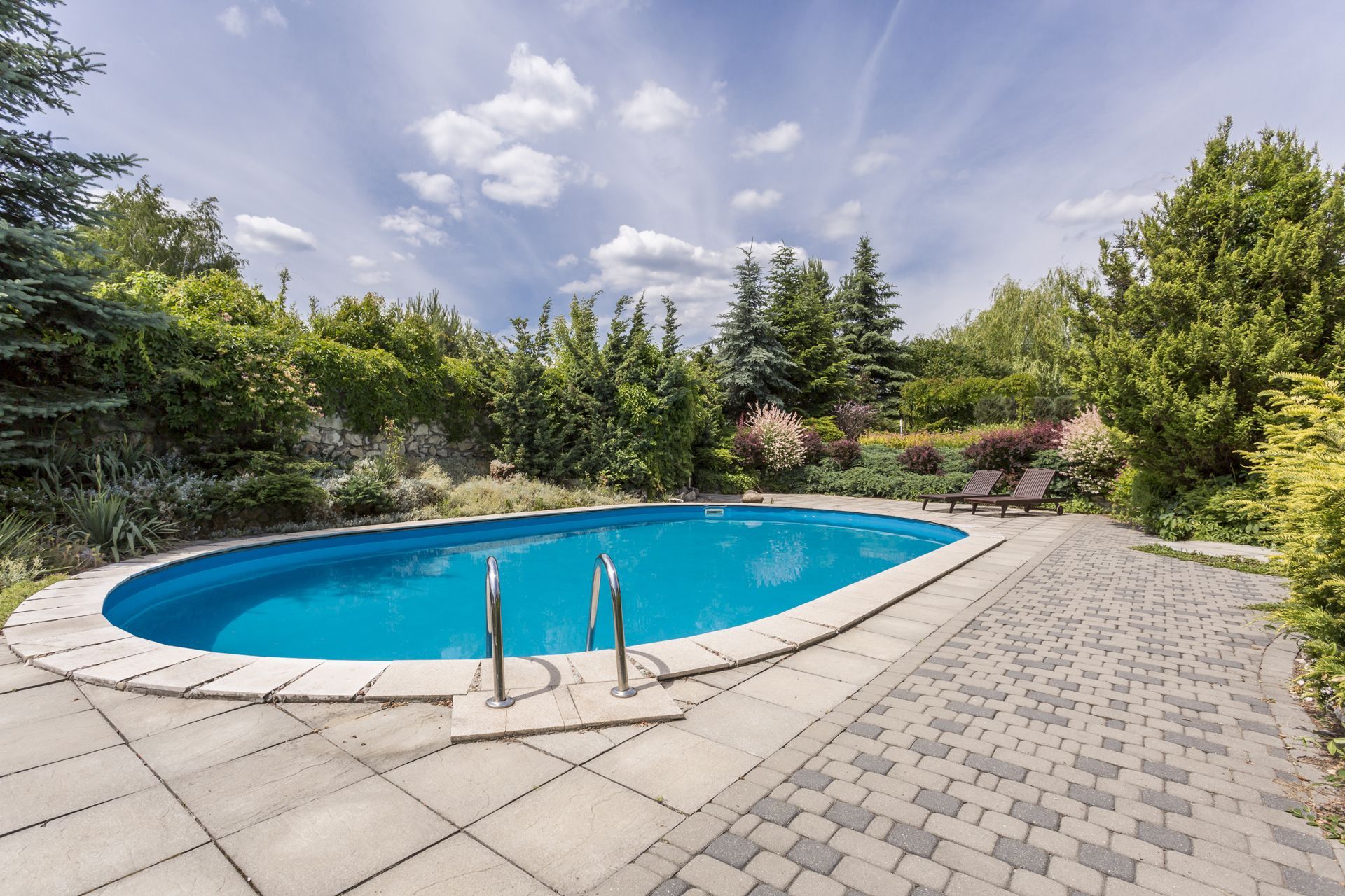 Oval-shaped pool surrounded by stone patio and lush greenery under a partly cloudy sky.