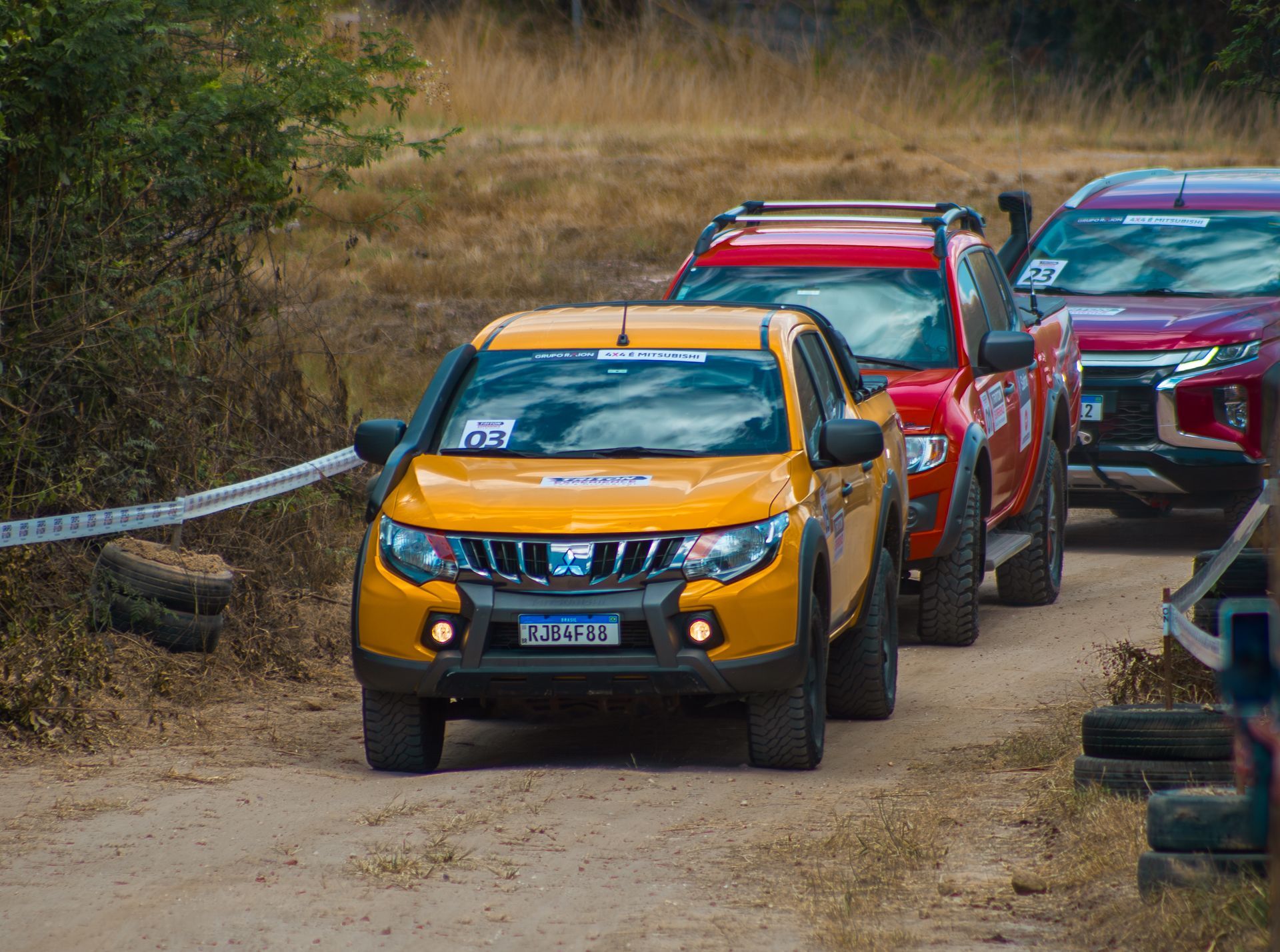 Três caminhonetes Mitsubishi off-road, nas cores amarela, vermelha e azul, trafegando em fila numa pista de terra.