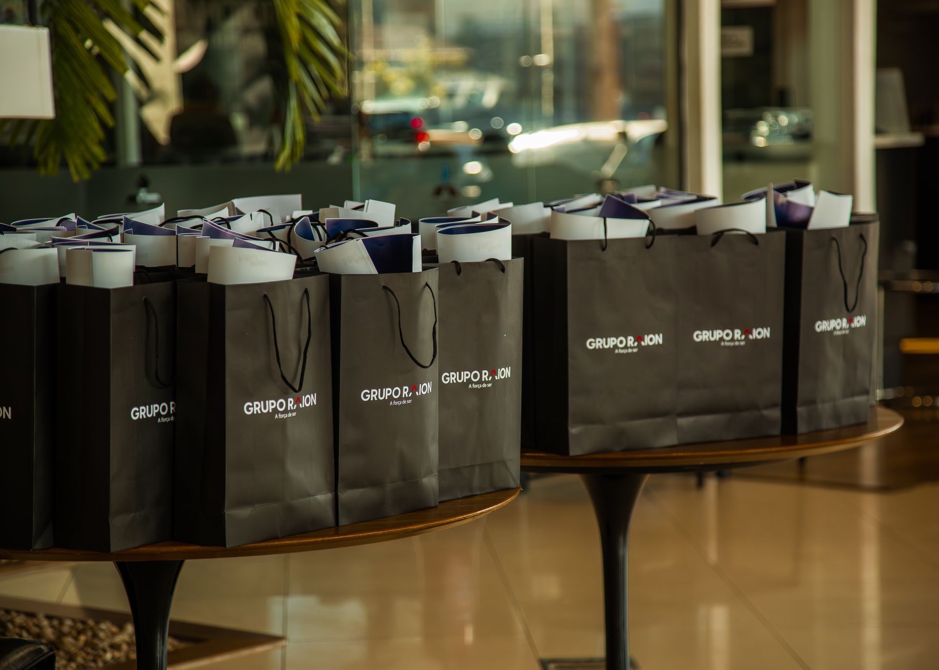Uma fileira de sacolas de presente pretas com logotipos brancos dispostas sobre uma mesa redonda em um ambiente interno.