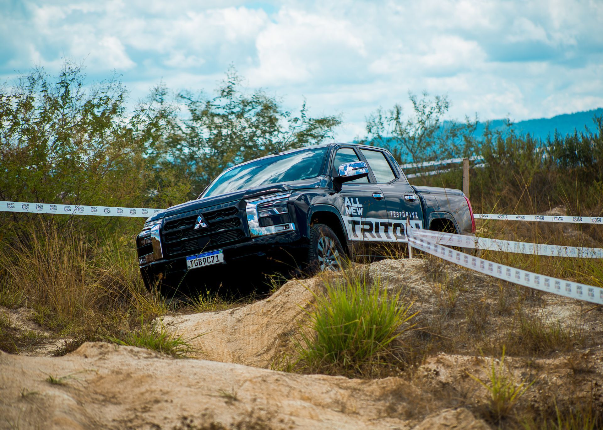 Uma picape Mitsubishi Triton escura trafega por um percurso off-road acidentado e arenoso, demarcado com fita branca.