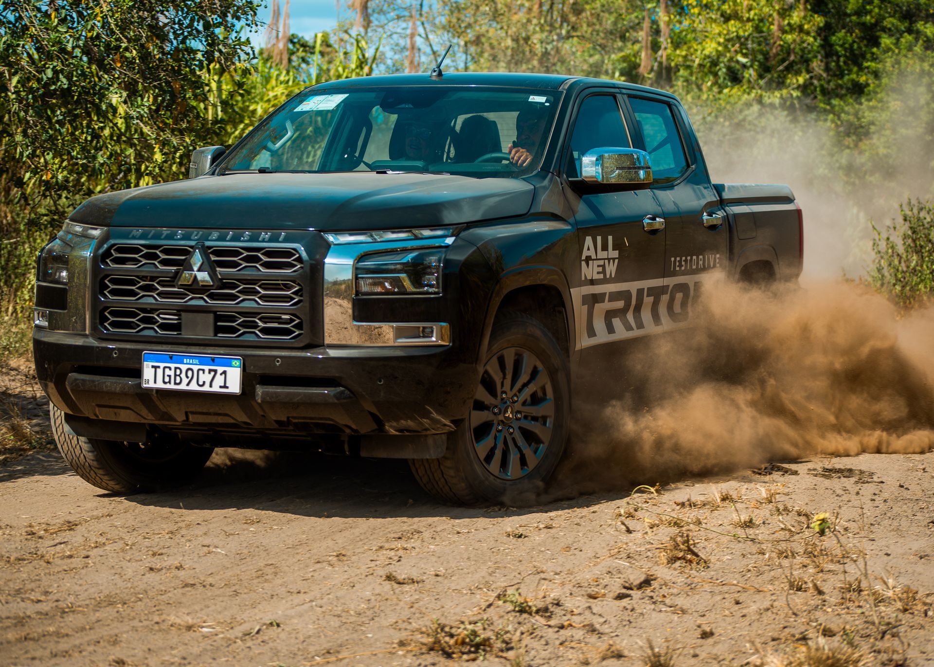 Uma caminhonete Mitsubishi Triton preta trafega por uma estrada de terra, levantando uma grande nuvem de poeira.