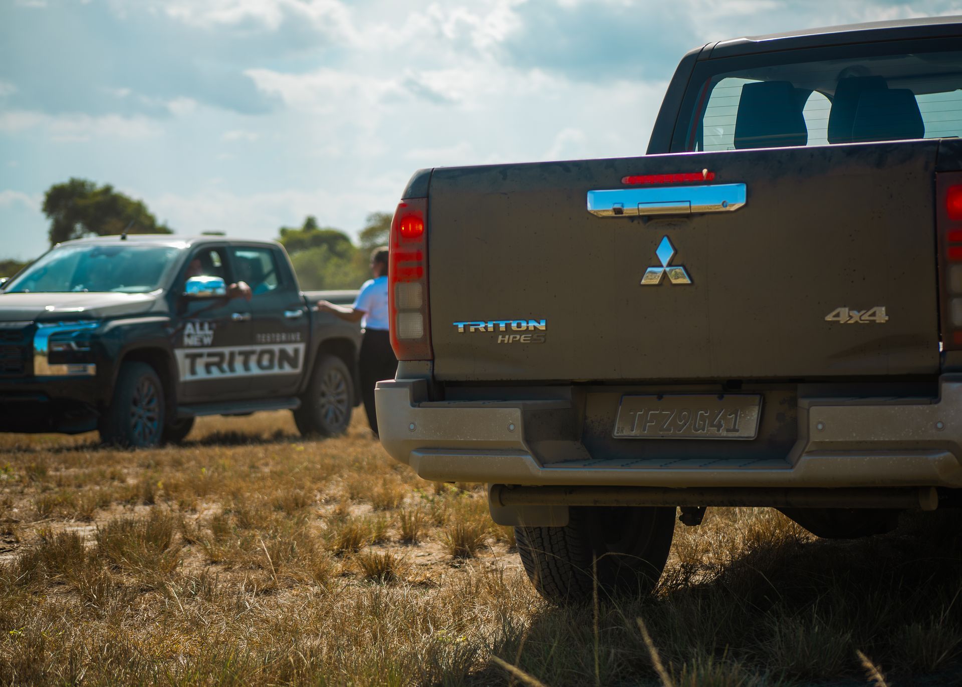 Duas caminhonetes Mitsubishi Triton estacionadas em um campo gramado e ensolarado.