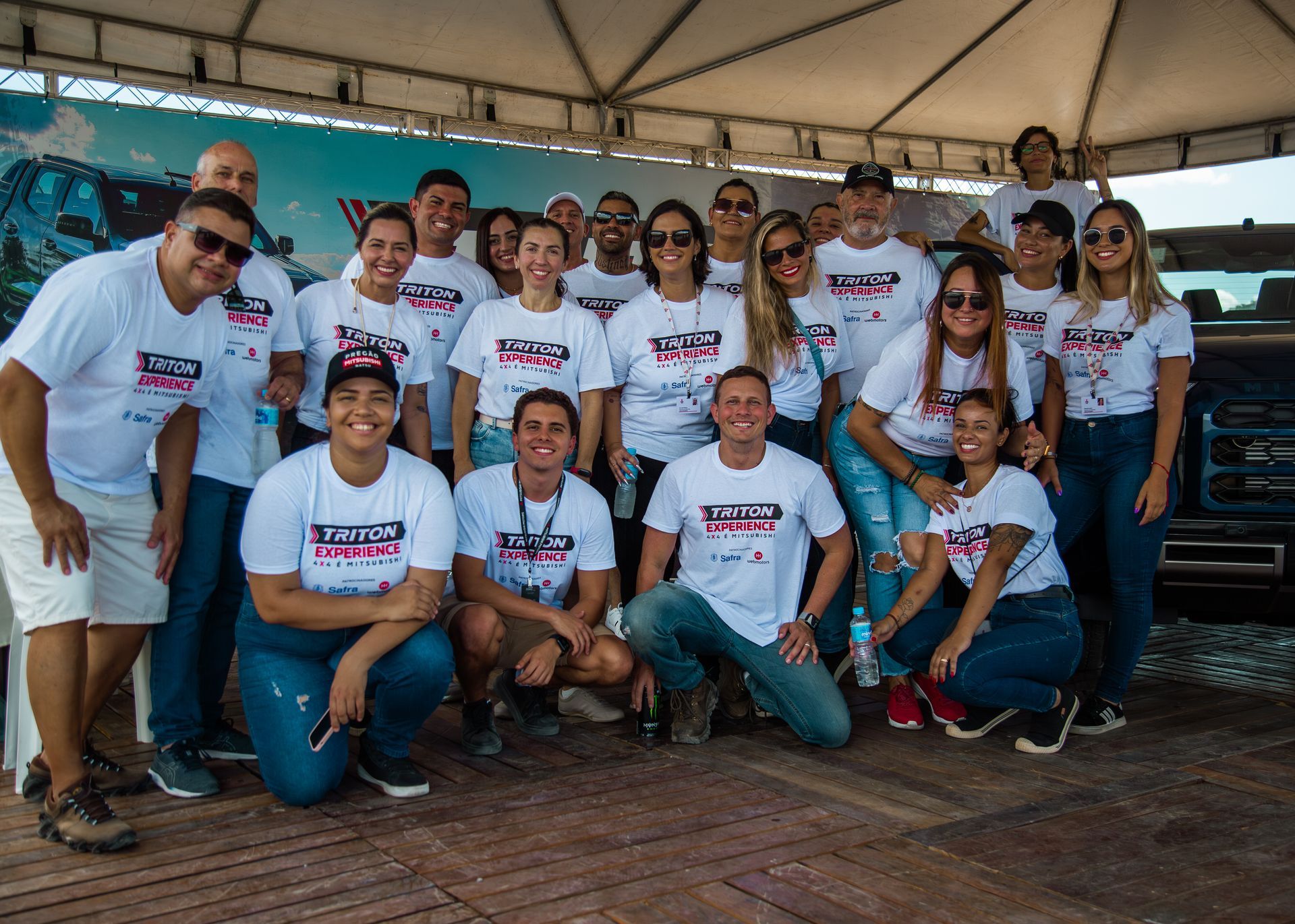 Um grupo de pessoas vestindo camisetas brancas iguais, usadas no evento, posa para uma fotografia sob uma tenda.