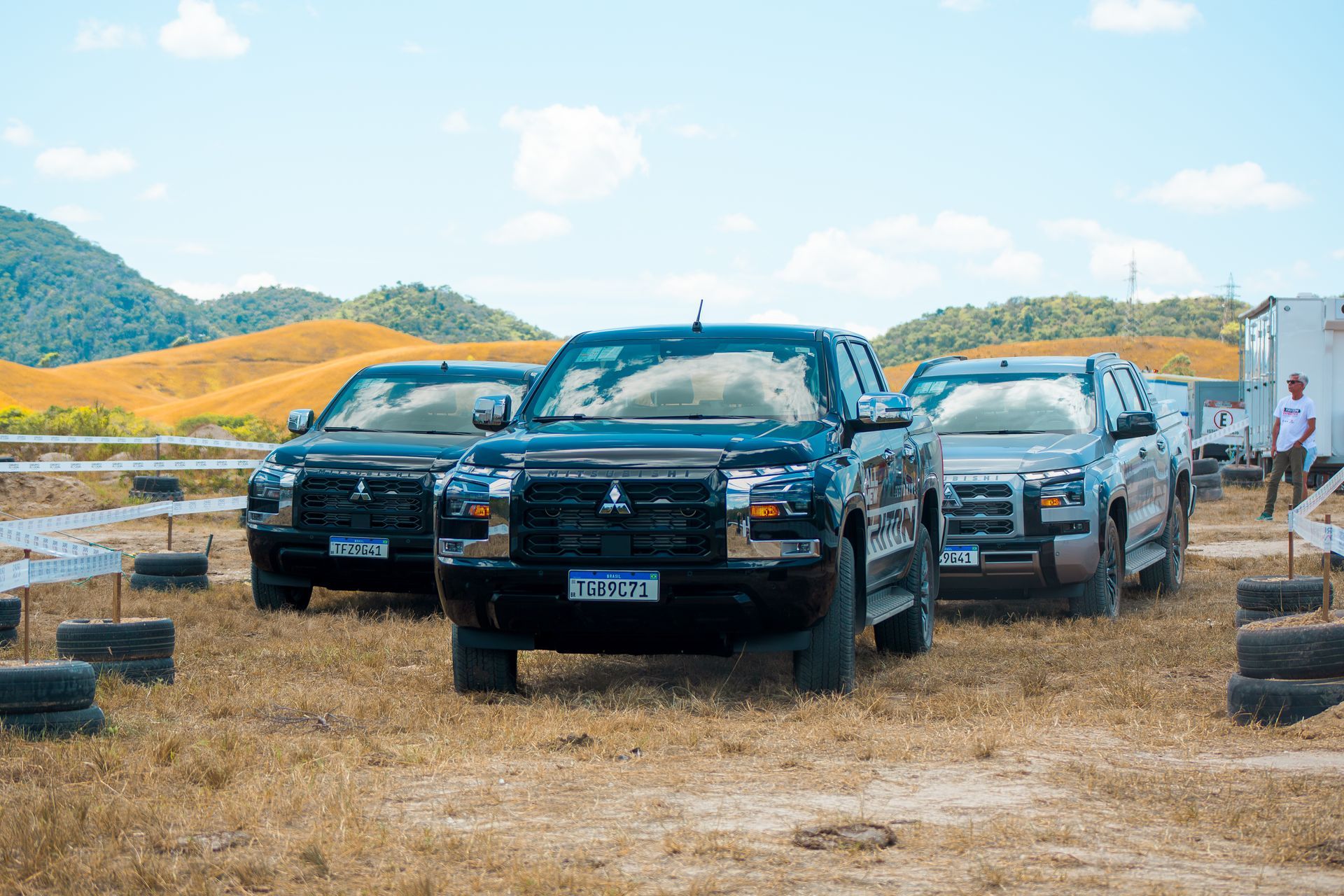 Três caminhonetes estacionadas em um campo gramado e seco, com colinas onduladas e uma pessoa em pé ao fundo.