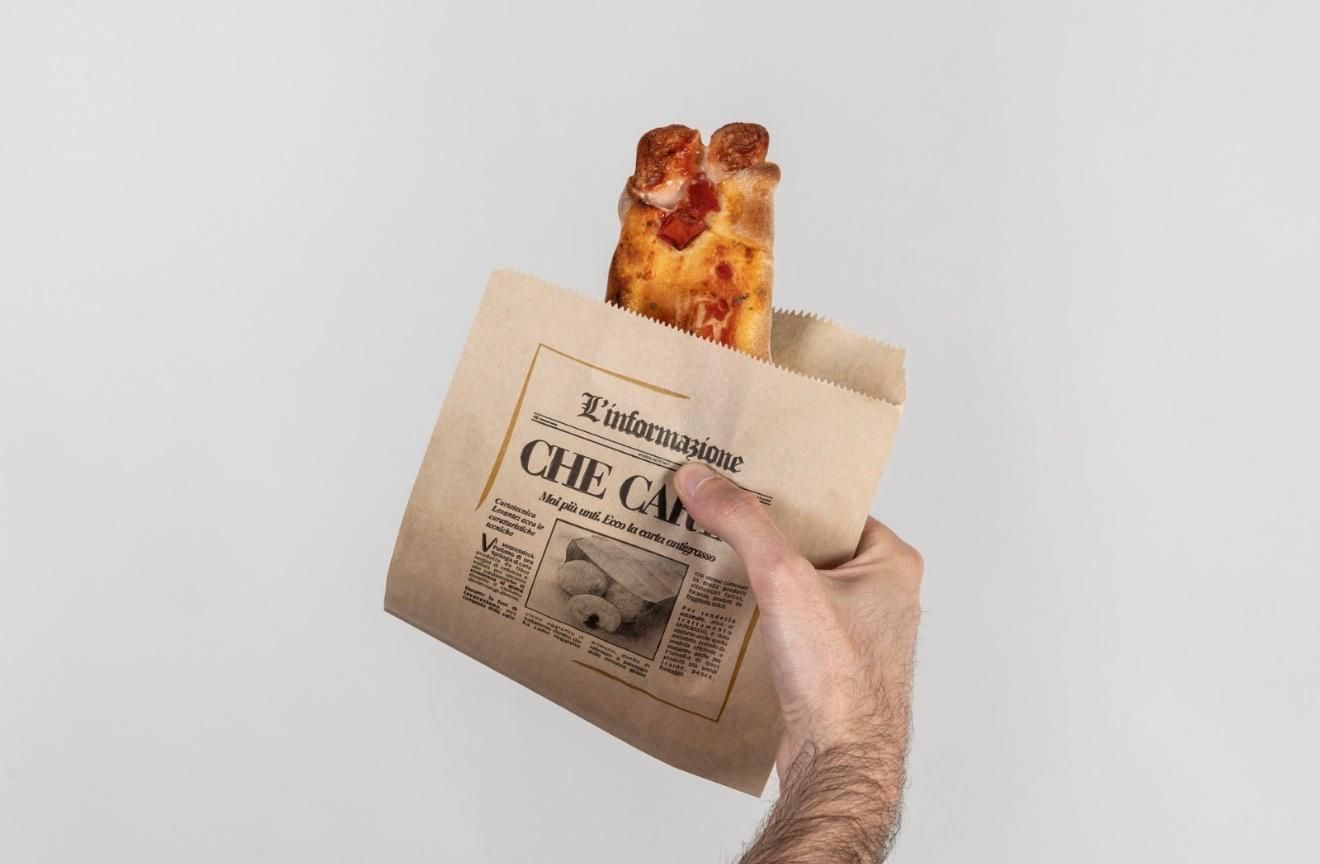 Mano che regge un sacchetto di carta con una fetta di pizza al salame piccante, su uno sfondo grigio uniforme.