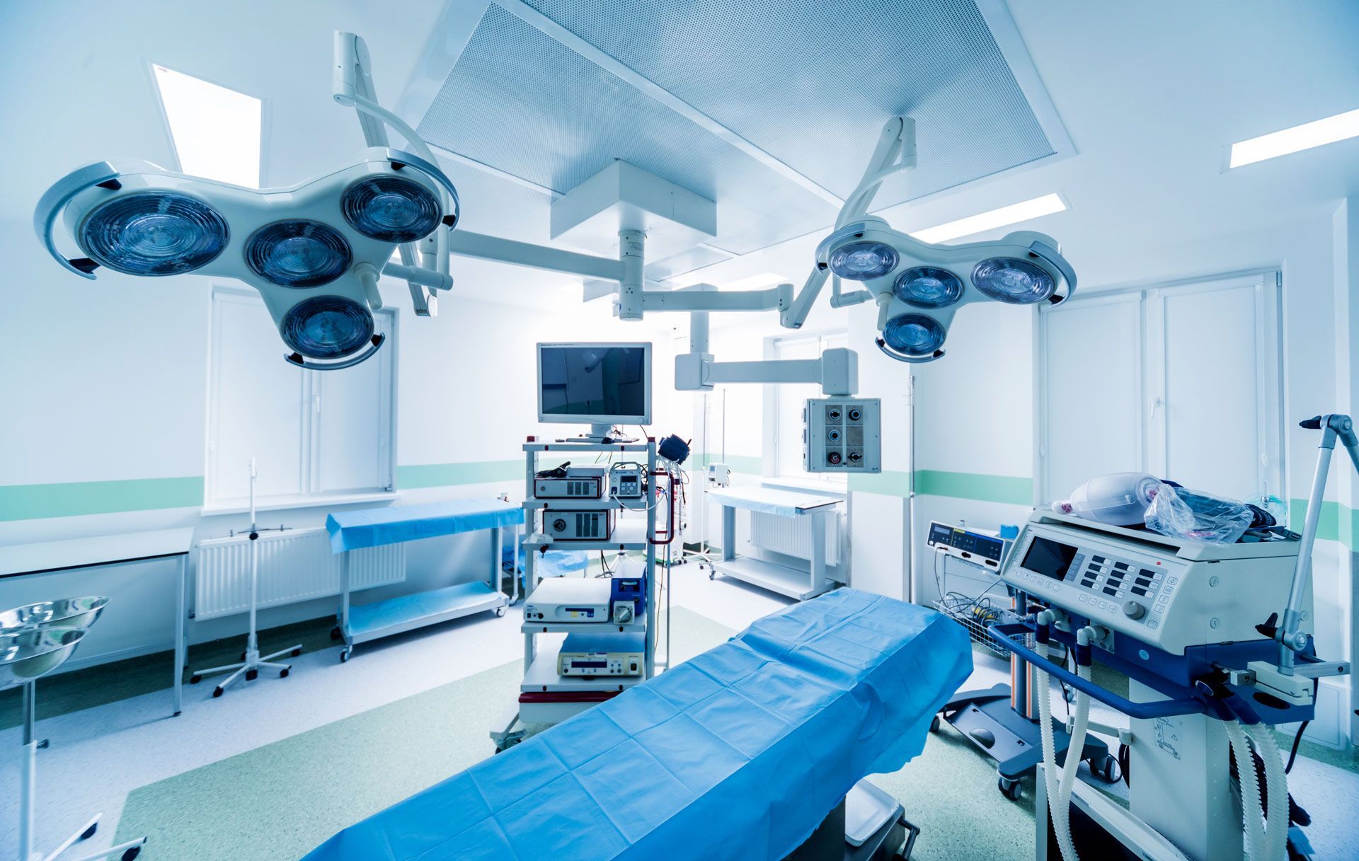 An operating room, with surgical lights, equipment, and a blue-covered patient bed, sterile and ready for a procedure.