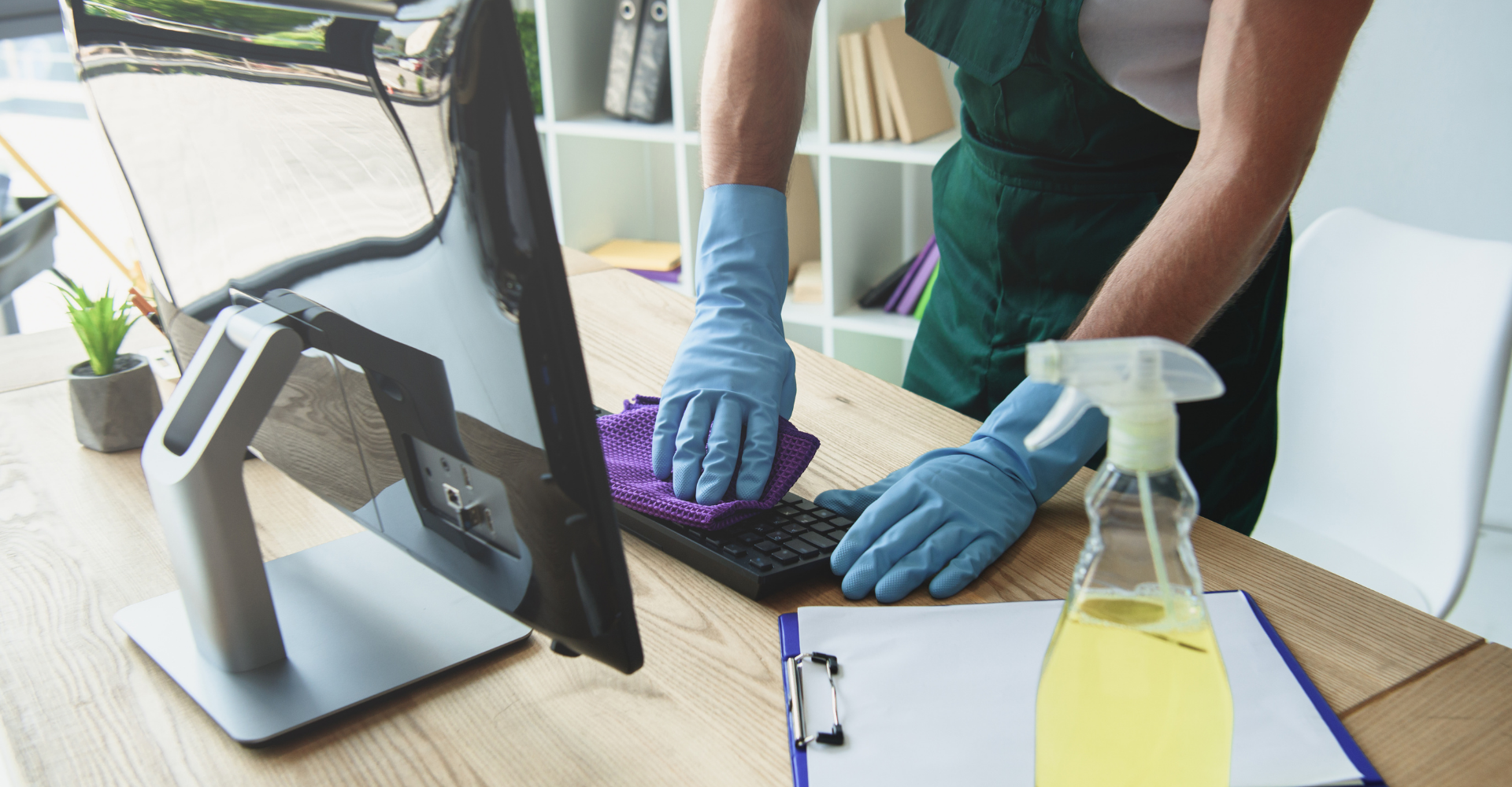 Commercial cleaner wiping down the computer keyboard set atop a wood grained desk