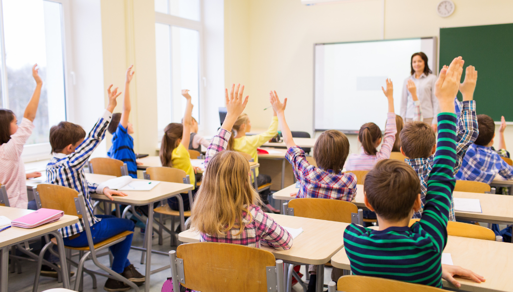 group of school kids with teacher sitting in classroom and raising hands