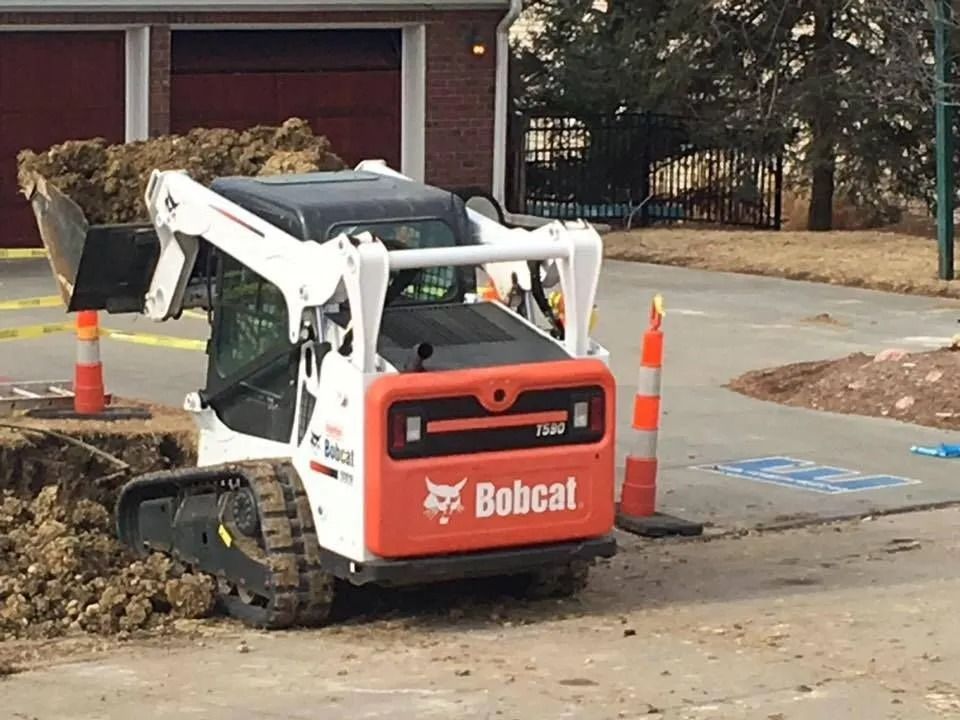 Bobcat skid-steer loader beside a dug-up curb with traffic cones in a driveway area