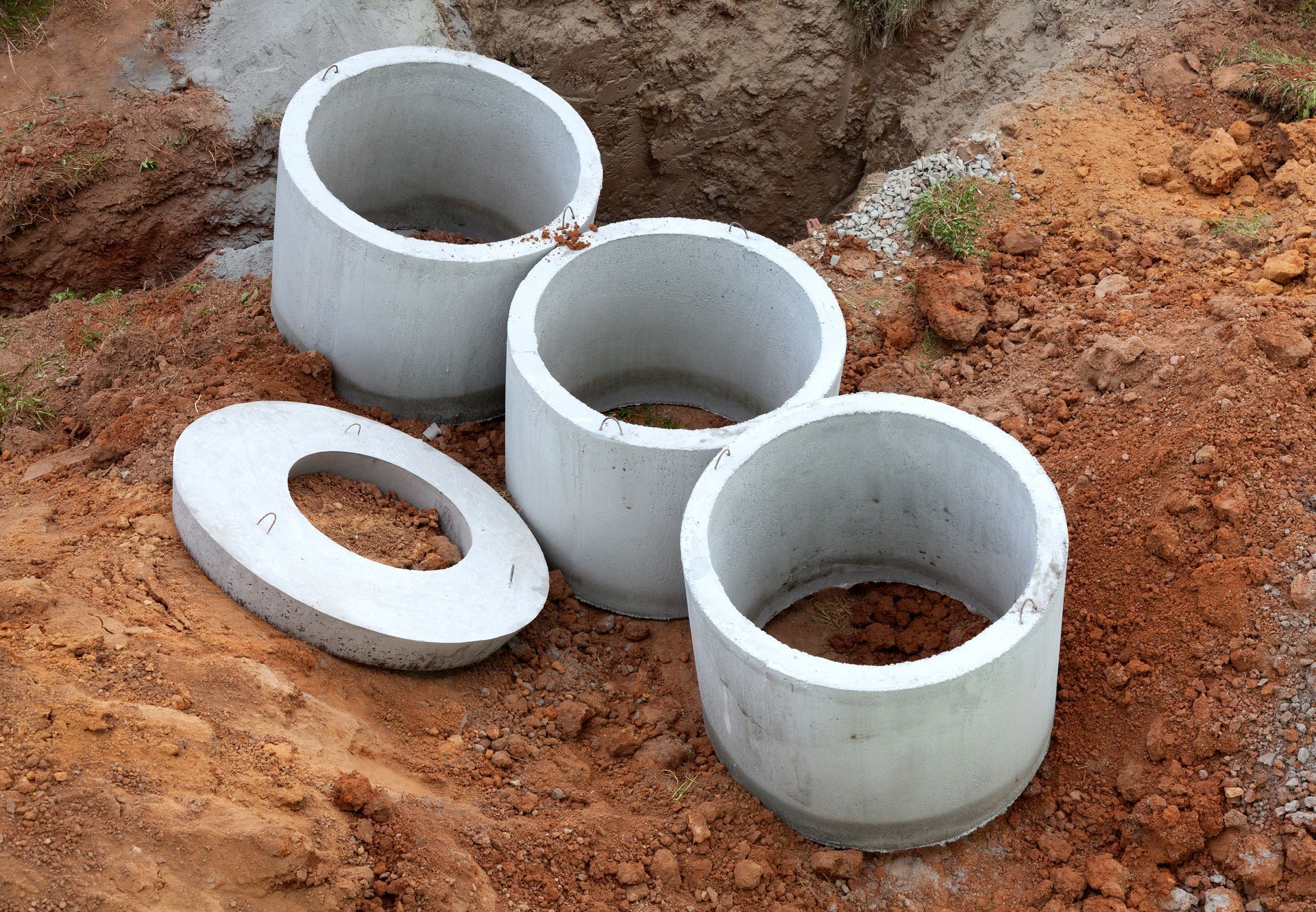 Three white concrete rings and a lid lying on reddish dirt in a construction area