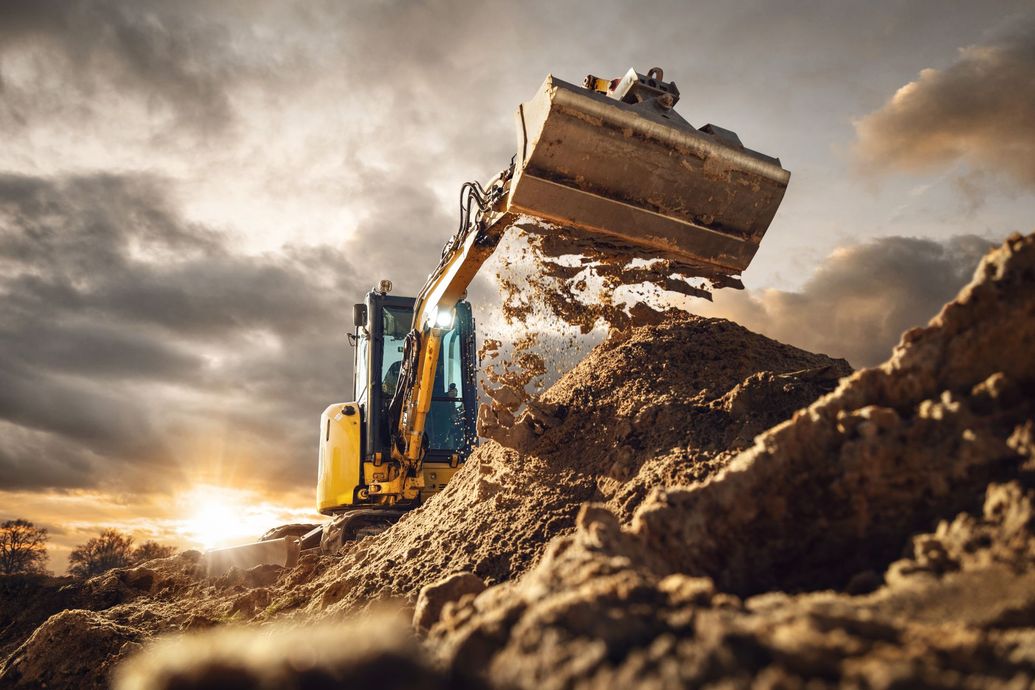 Yellow excavator dumping dirt on a large earth mound under a cloudy sky at sunset