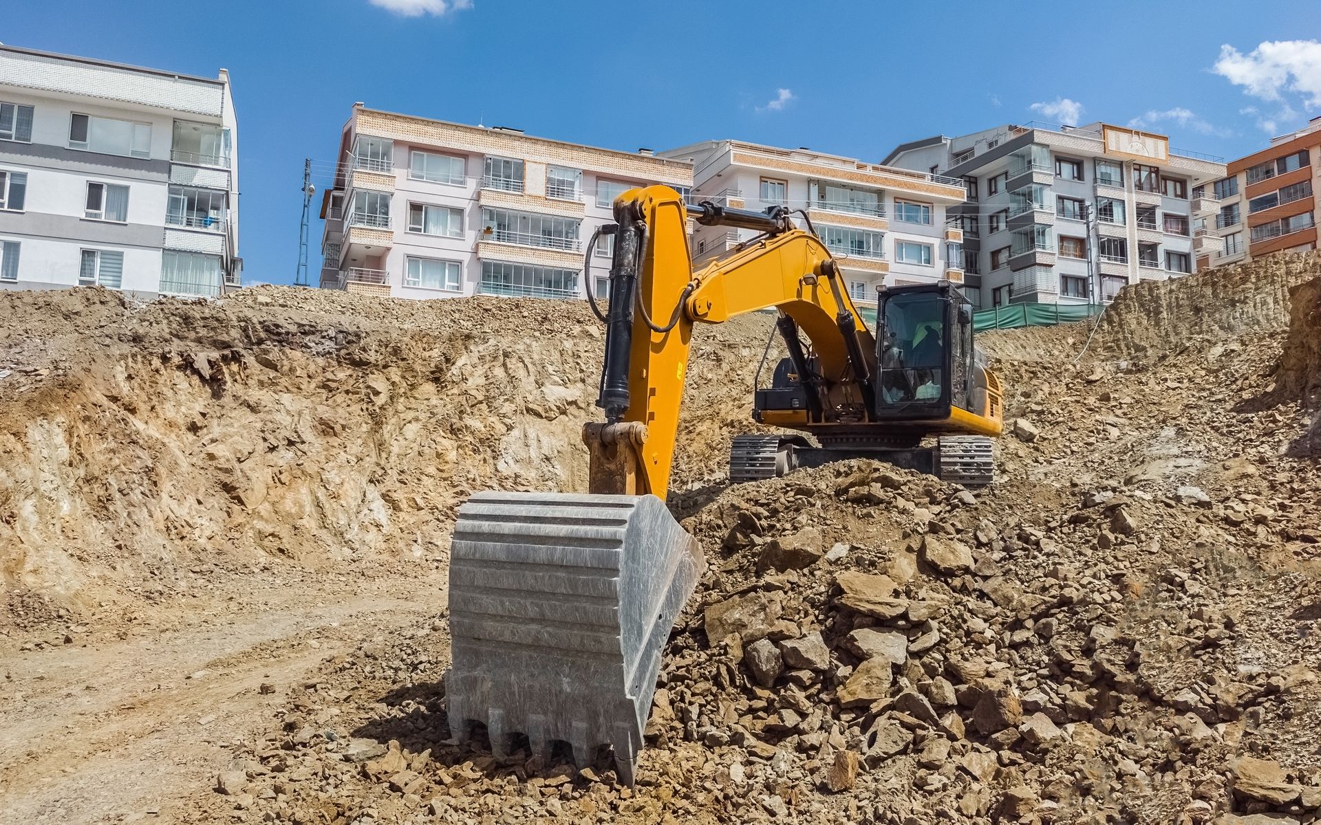 Yellow excavator digging in a construction site with apartment buildings in the background