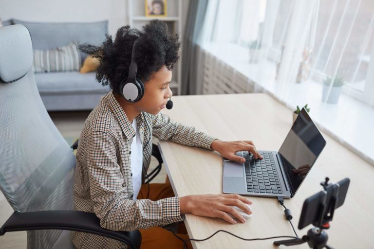 Person with headphones, at laptop, recording video. Neutral setting with a window.