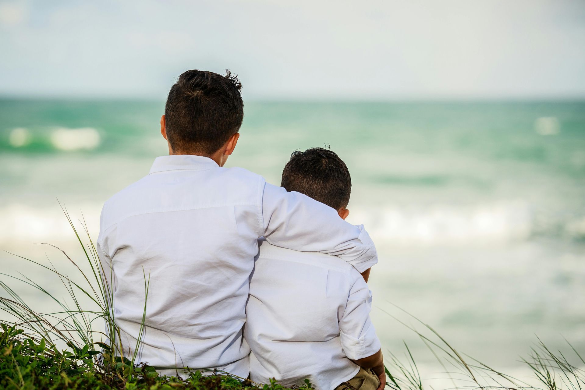 A father and son are sitting on the beach looking at the ocean.