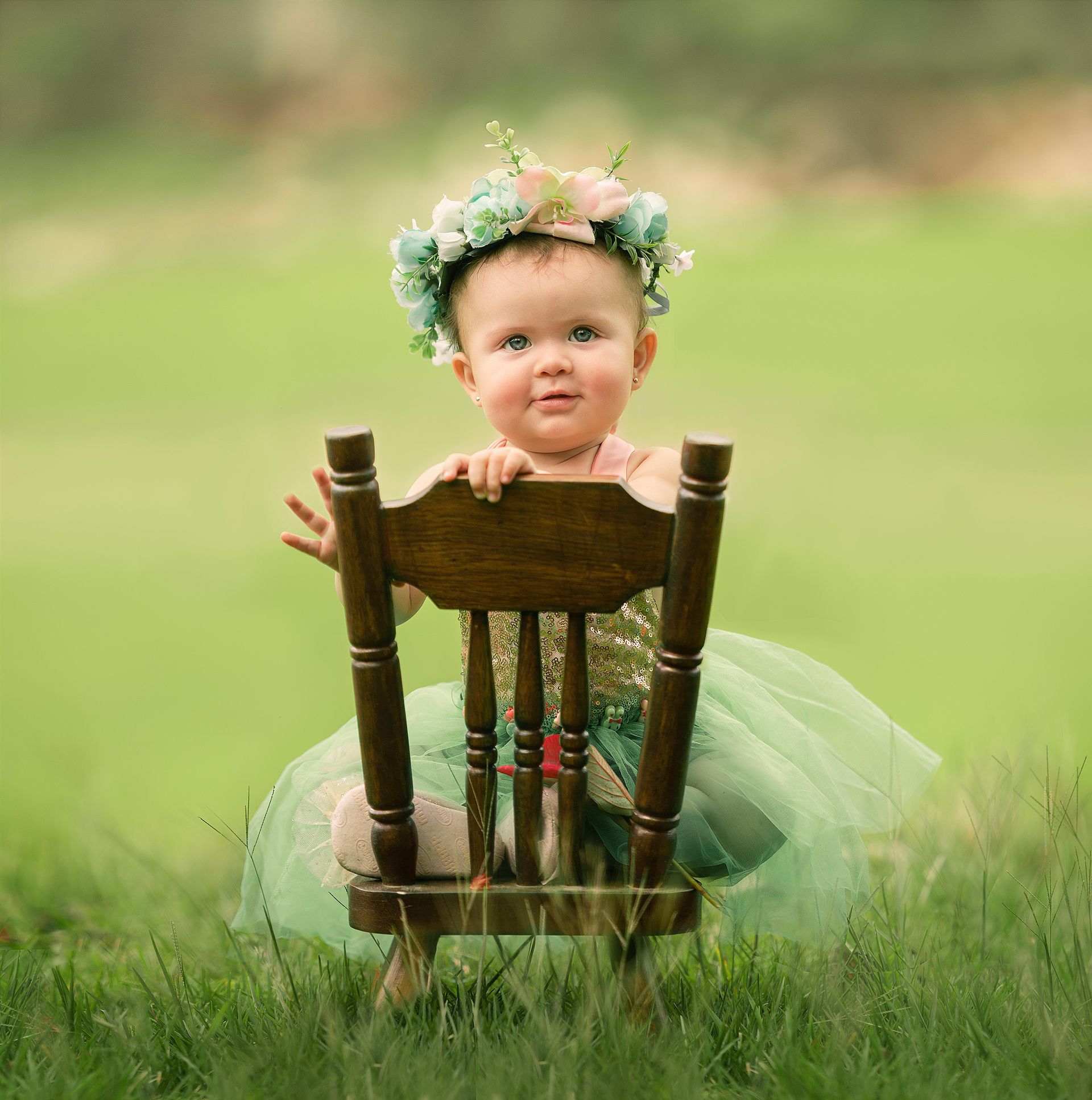 Baby wearing flower crown, sitting in a small wooden chair in a grassy field. The baby is smiling.