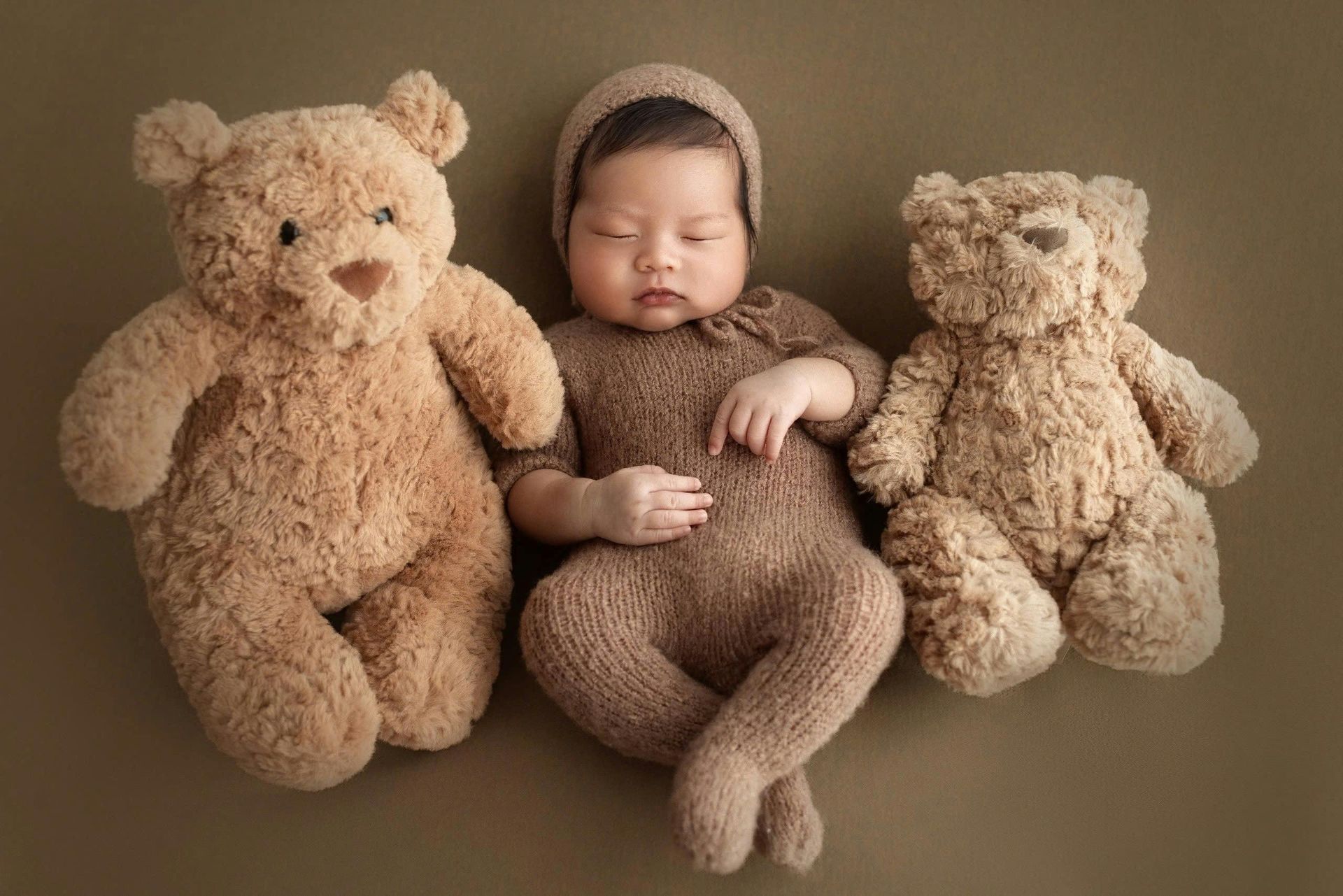 Newborn baby in a brown outfit and cap, sleeping between two teddy bears on a brown backdrop.
