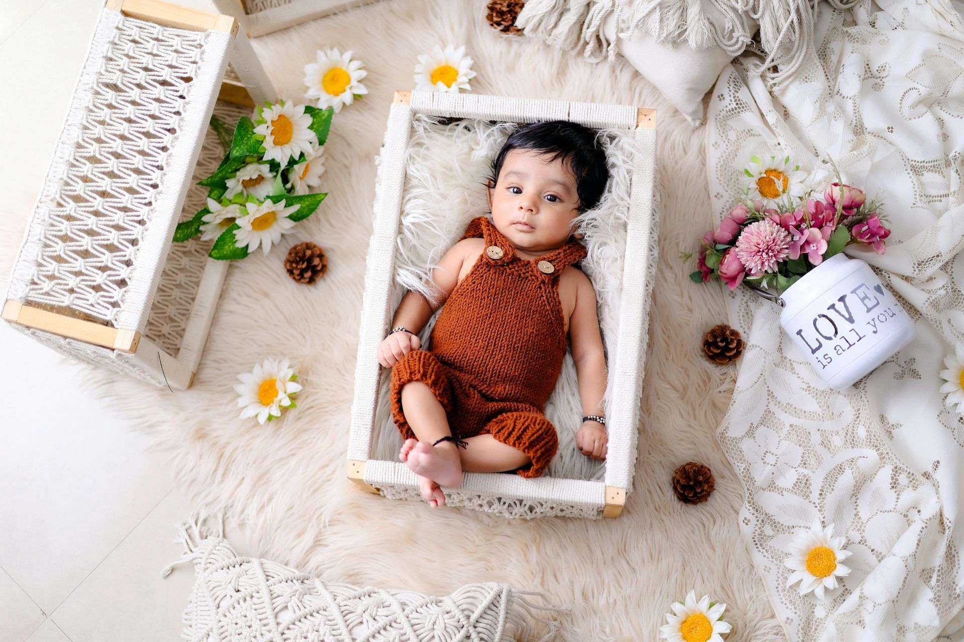 Baby in a brown knitted jumpsuit, lying in a white woven basket, surrounded by flowers and dรฉcor.from Australian Nanny Agency Website