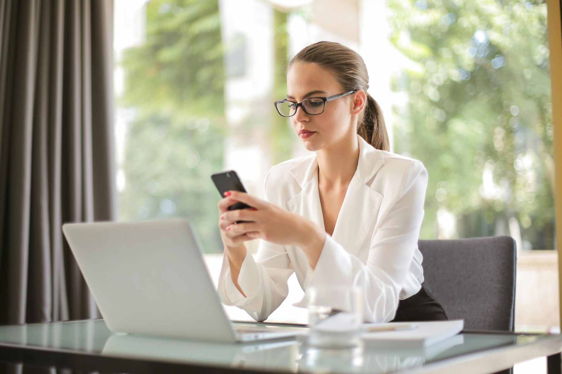 Woman in glasses and white blazer using phone, working on laptop at desk at Australian Nanny Agency