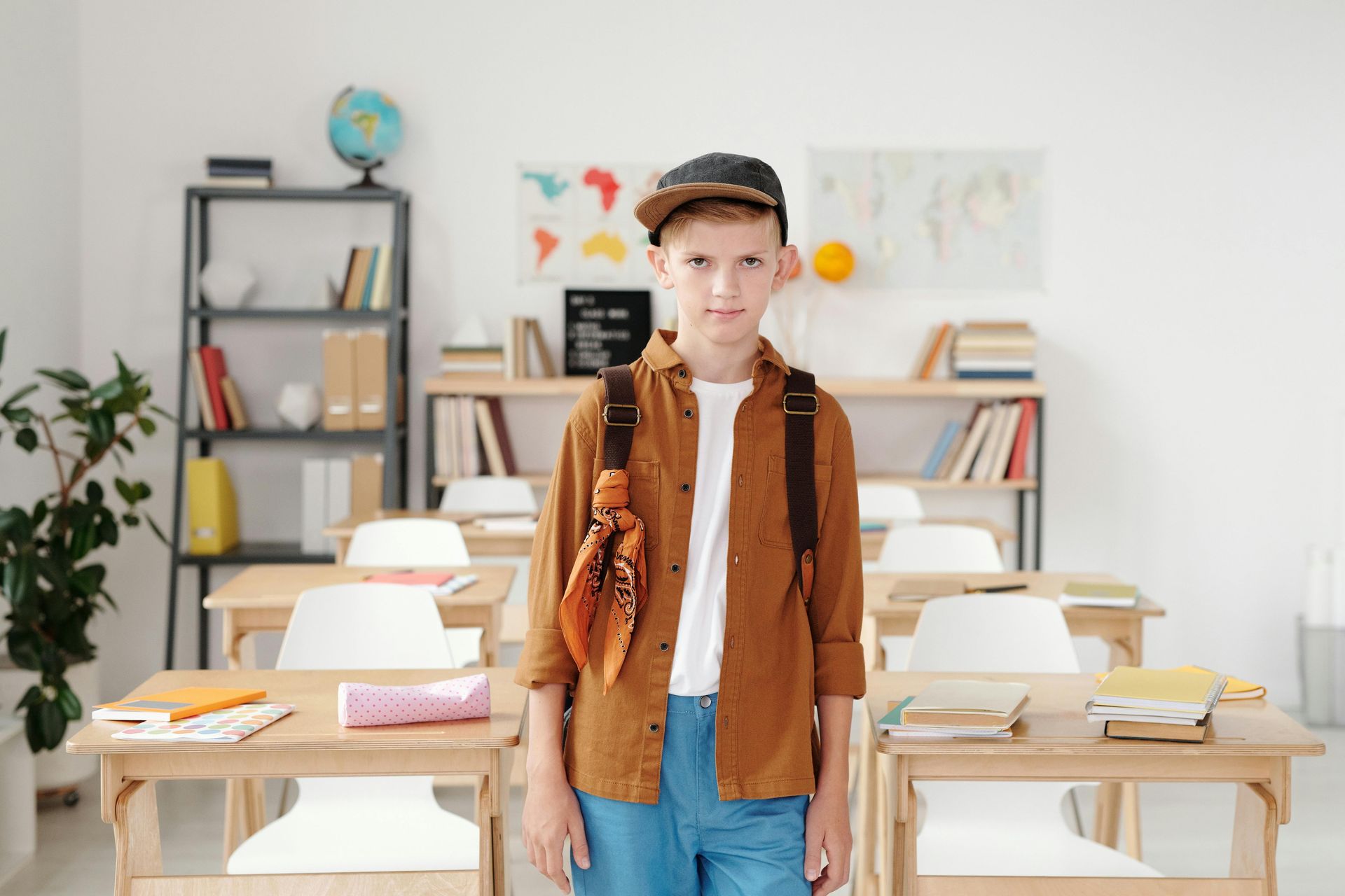 Boy in classroom wearing backpack, cap, and brown shirt, smiles in front of desks, books, and map.