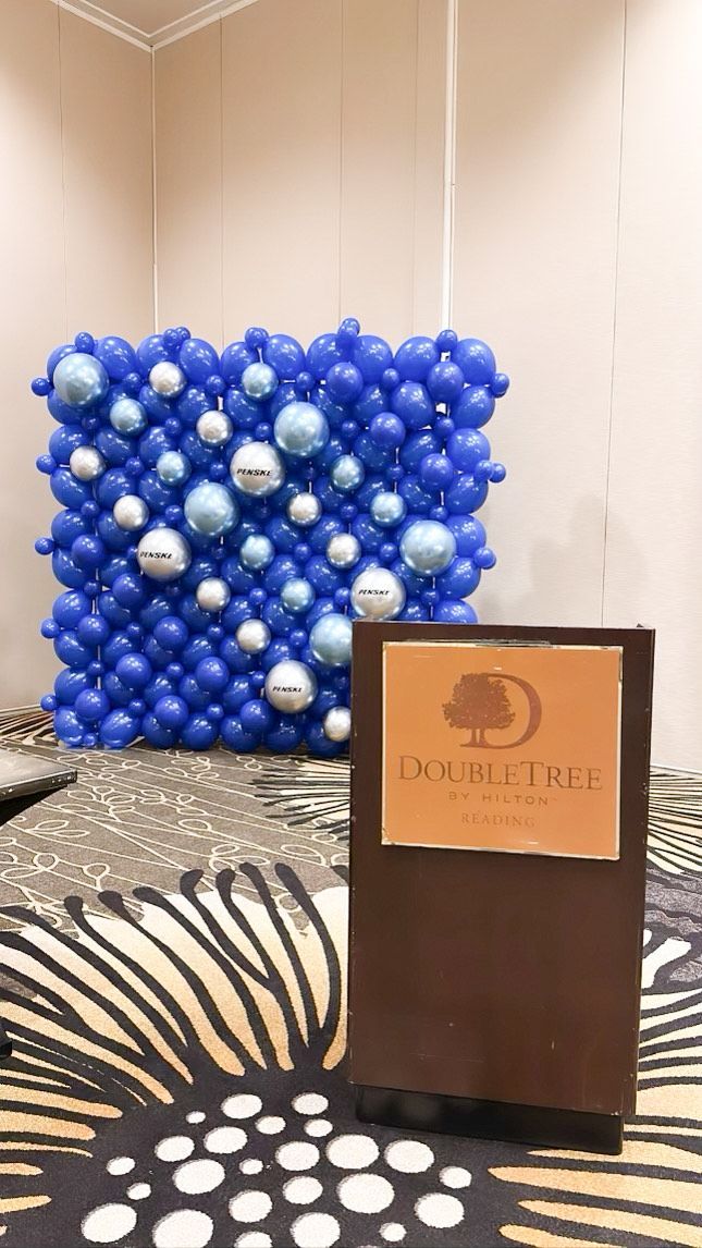 Blue and white balloon display behind a brown podium with a logo sign on a patterned carpet.