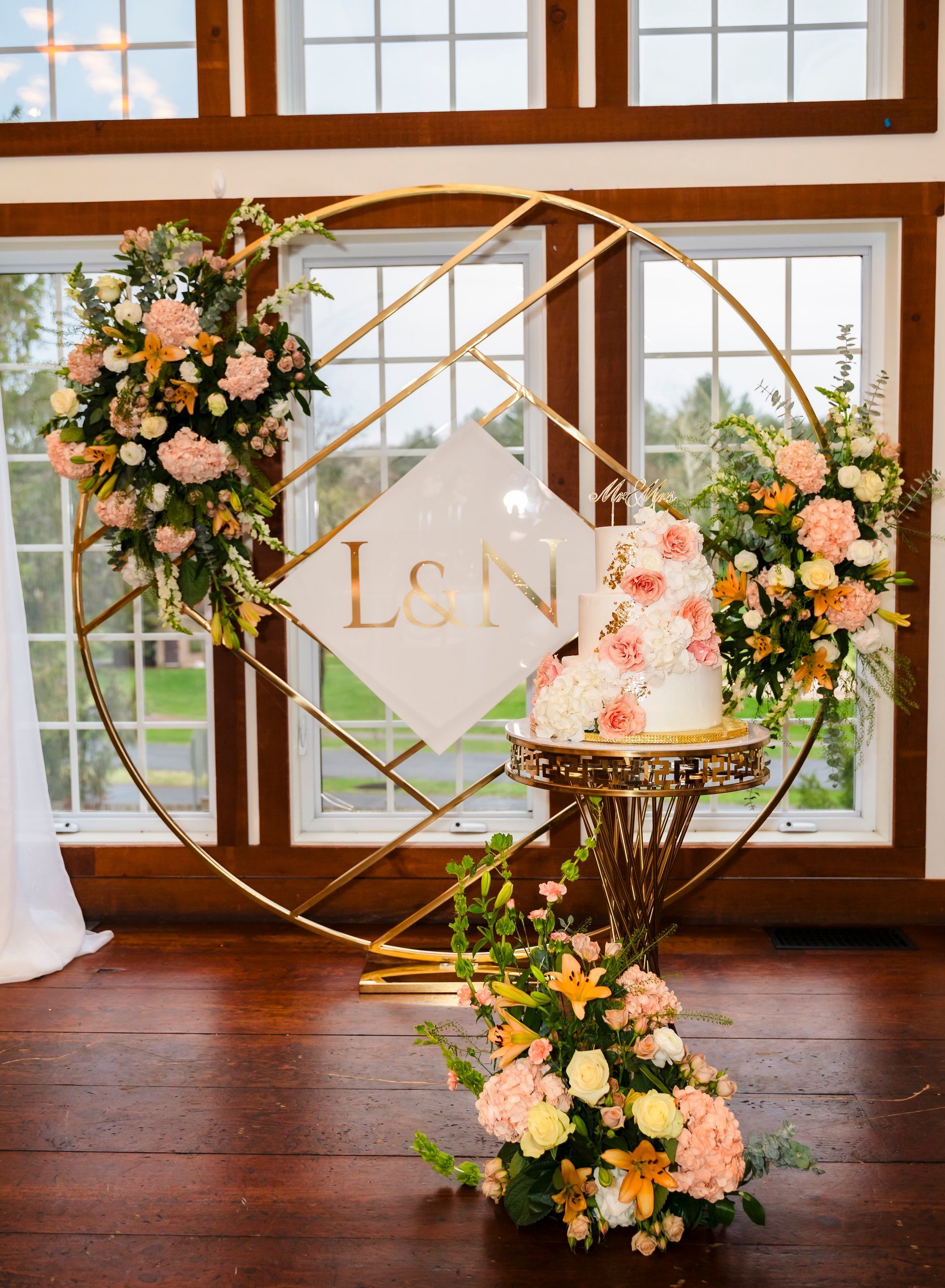 Wedding cake display with floral gold hoop backdrop and pastel flowers by a window