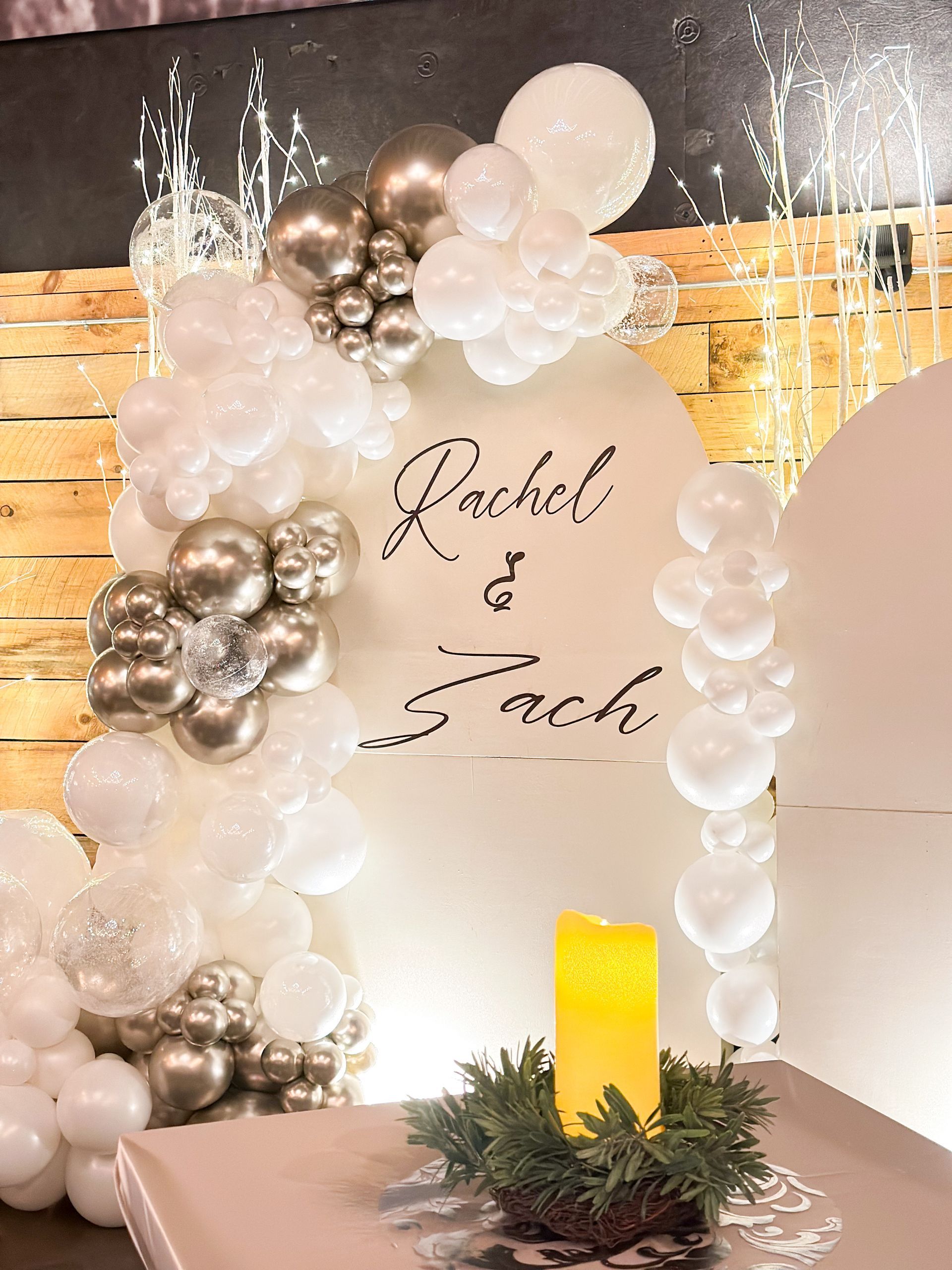 White and silver balloon arch with “Rachel & Zach” sign and candles on a festive backdrop
