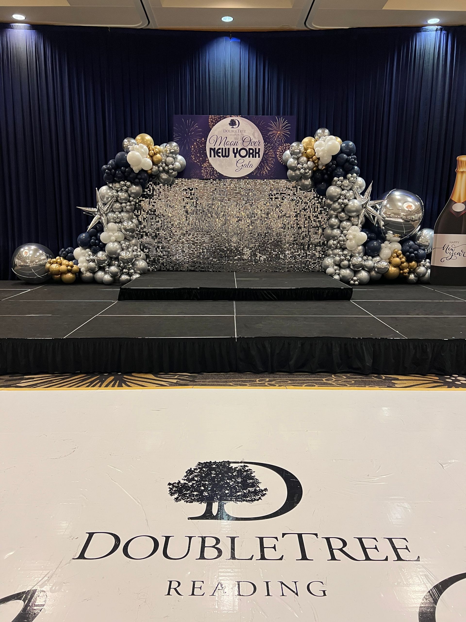 Decorated event stage with silver and navy balloons, shimmering backdrop, and DoubleTree by Hilton logo on the floor