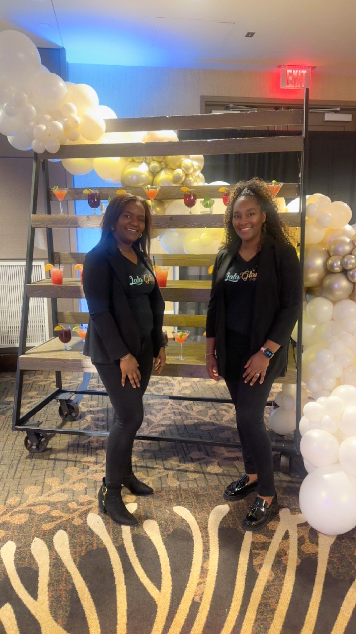 Two women in black outfits stand in a decorated room with white and gold balloons and shelves behind them.